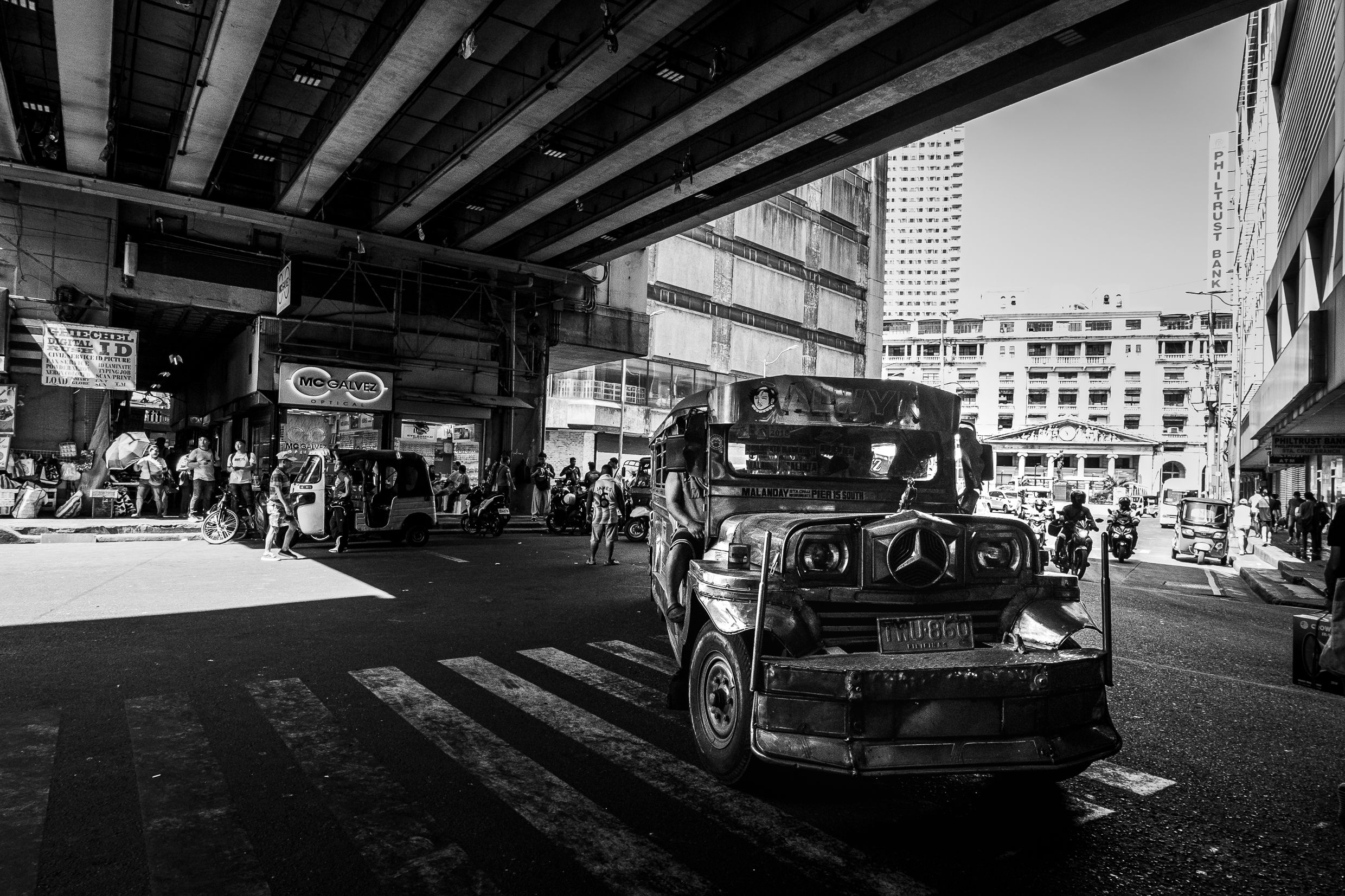 Black and white street view under a bridge in Manila with jeepneys and pedestrians.