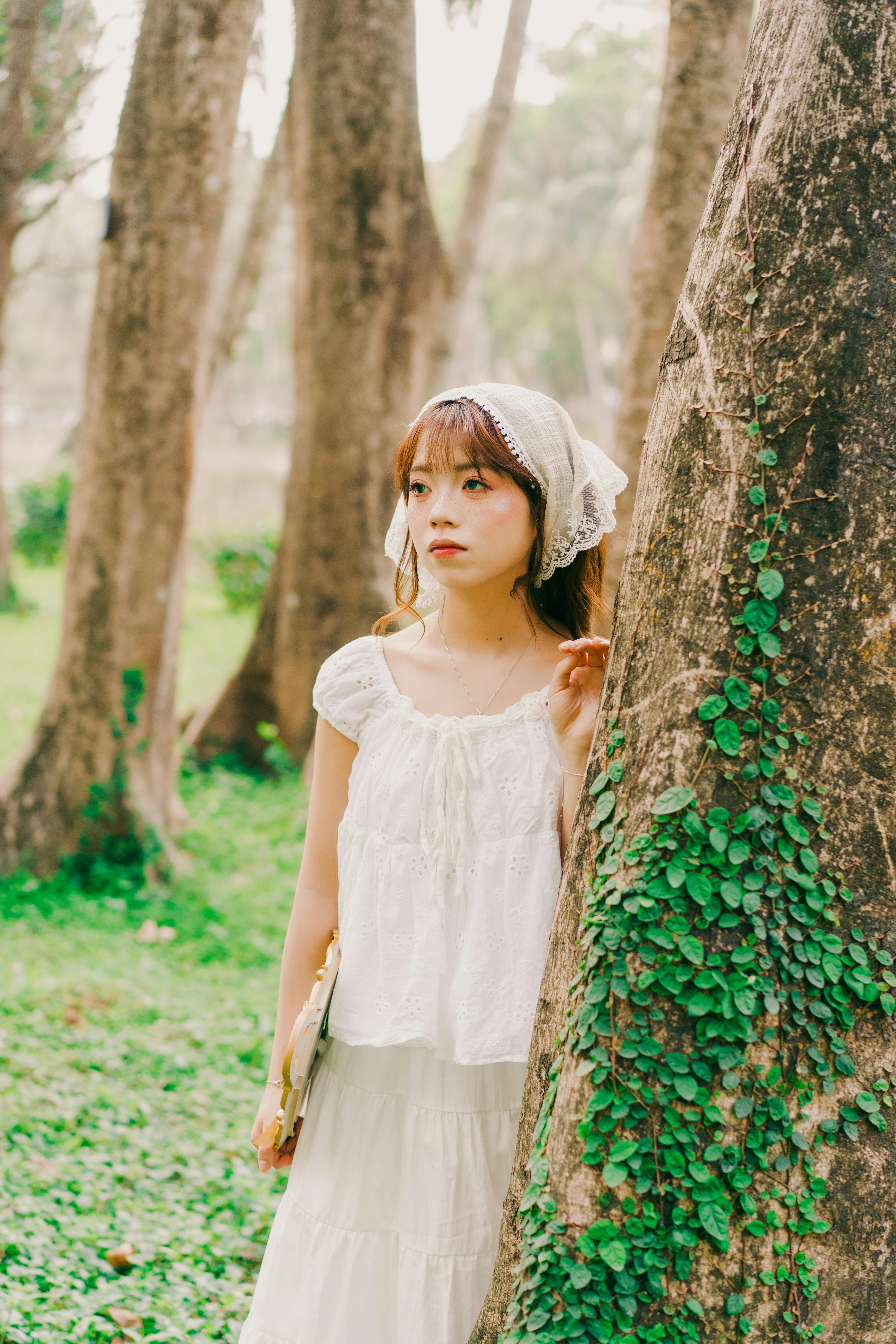 A young woman leans against a tree in a park in Hanoi, Vietnam, wearing a traditional outfit.
