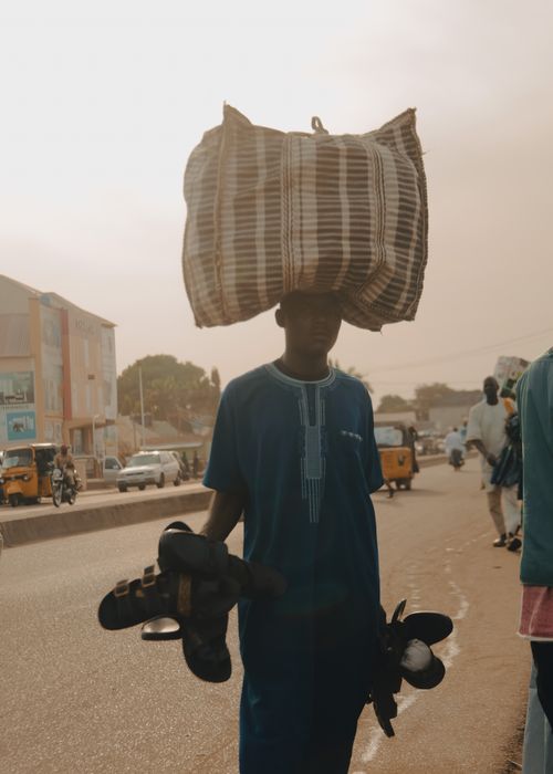 Free Man carrying bag on head and holding sandals in bustling street market scene. Stock Photo