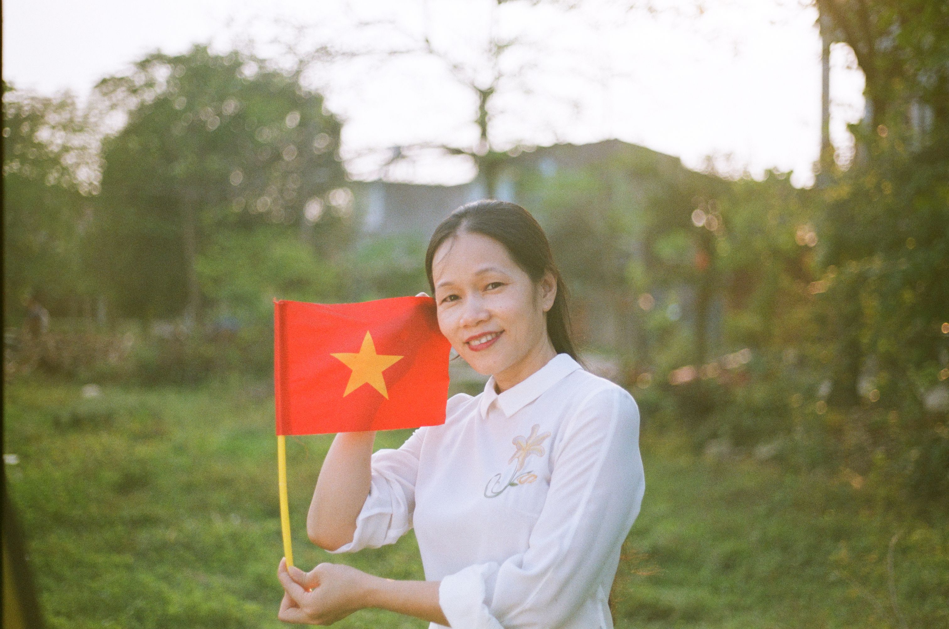 Smiling woman in white holding a Vietnamese flag in a sunny outdoor setting.