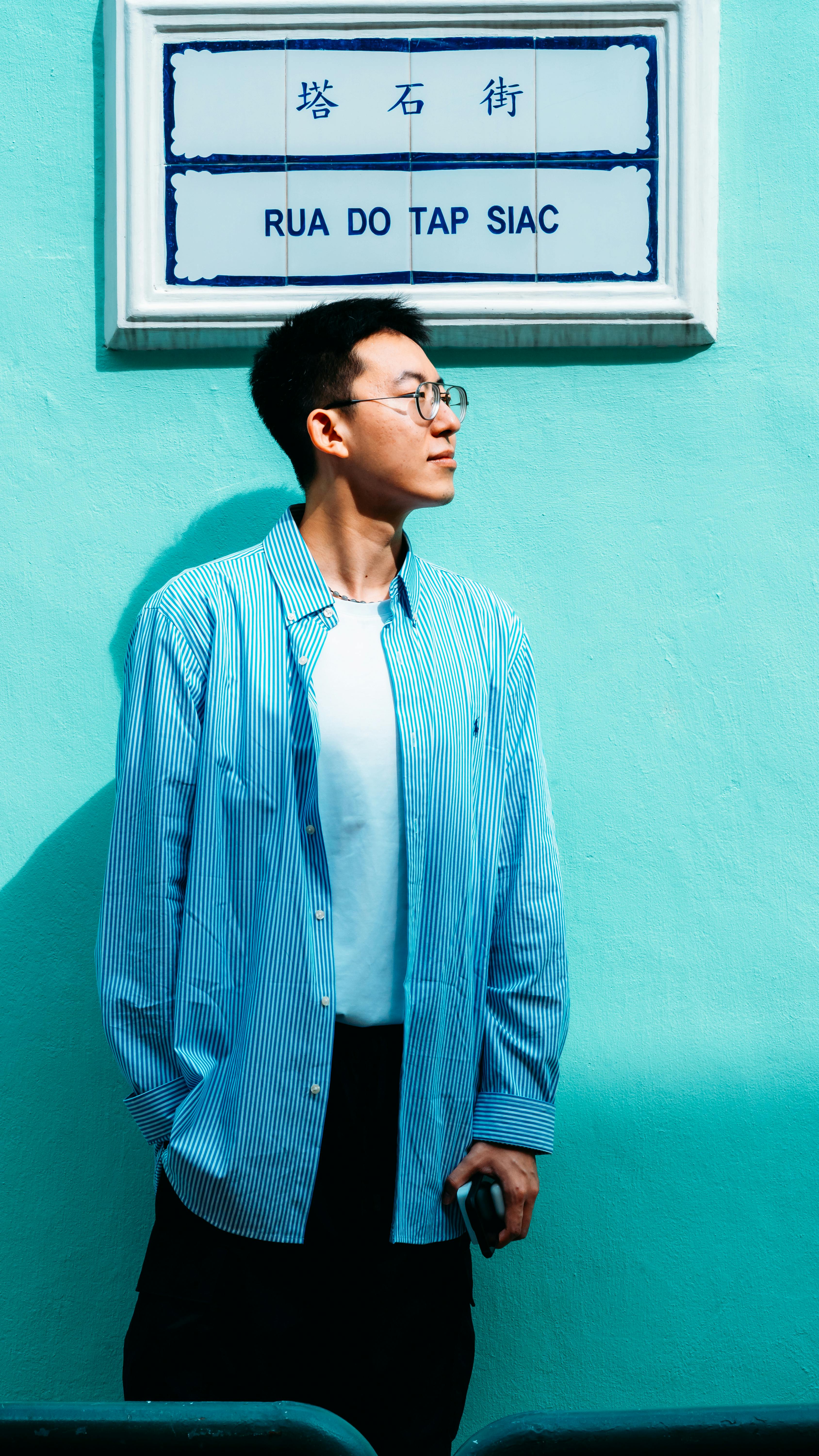 Young man stands against teal wall beside Rua do Tap Siac street sign.