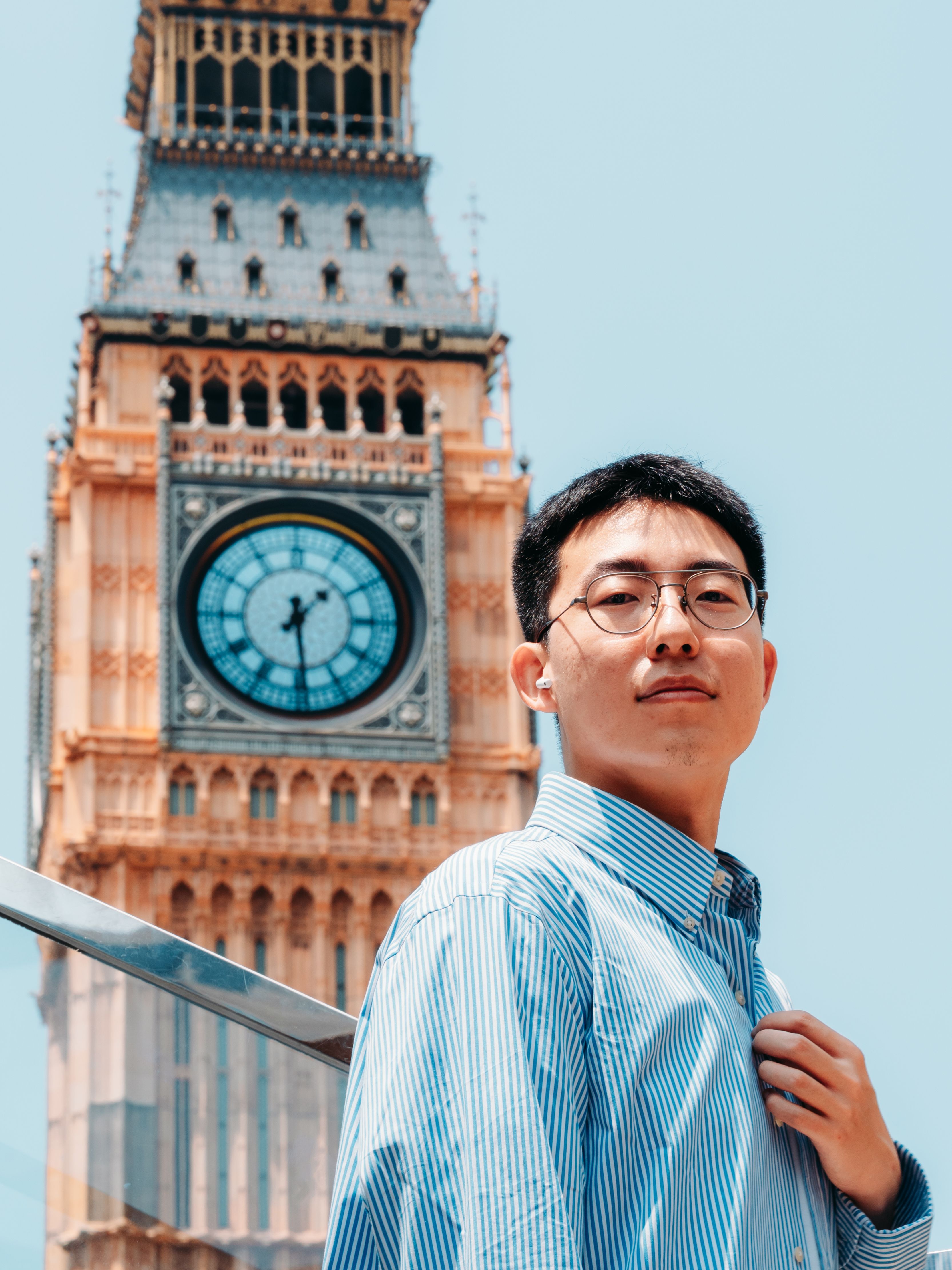 Confident young man in glasses standing before Big Ben under a clear sky.