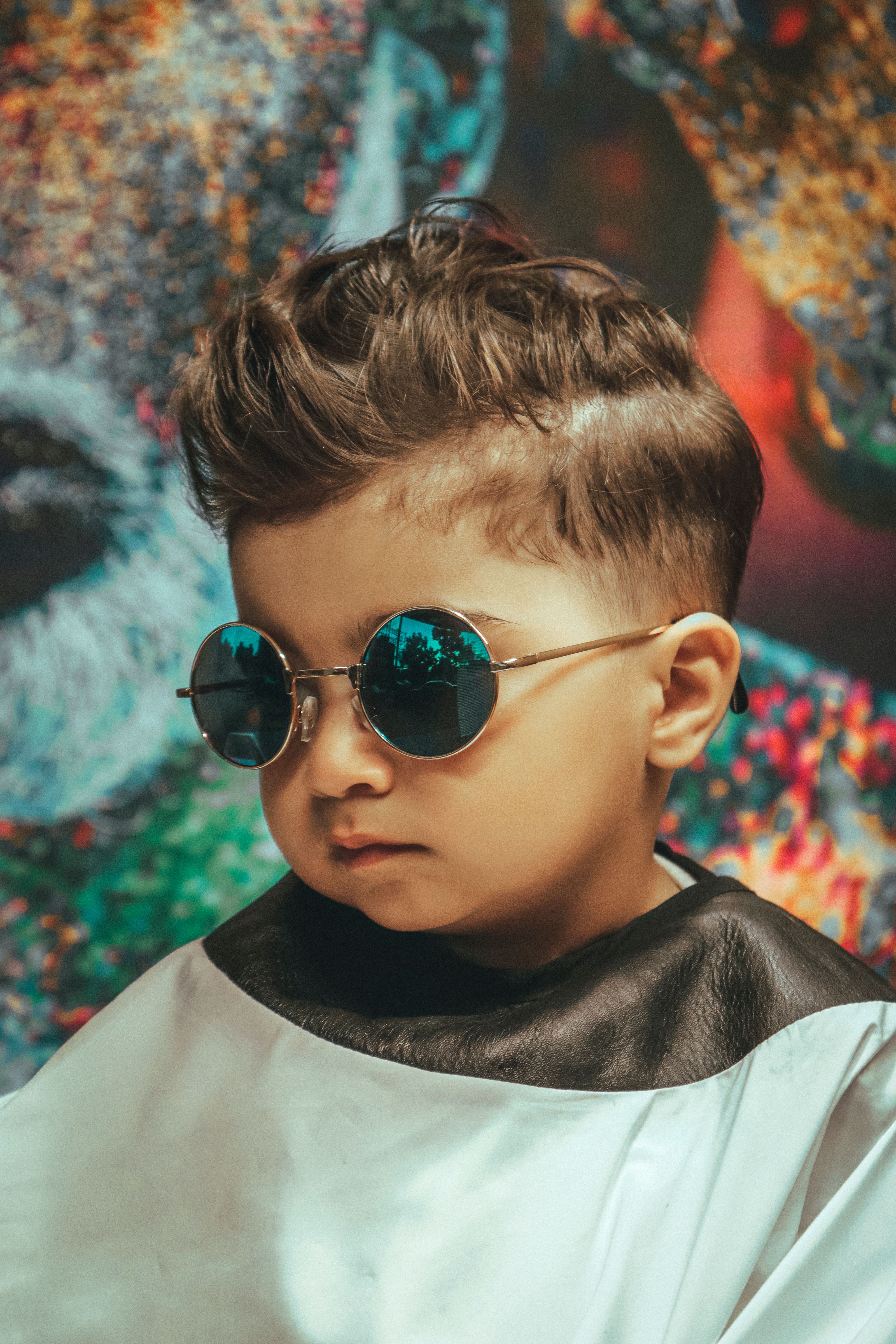 Criança jovem com um corte de cabelo moderno e óculos escuros, posando em um ambiente interno contra um fundo artístico.