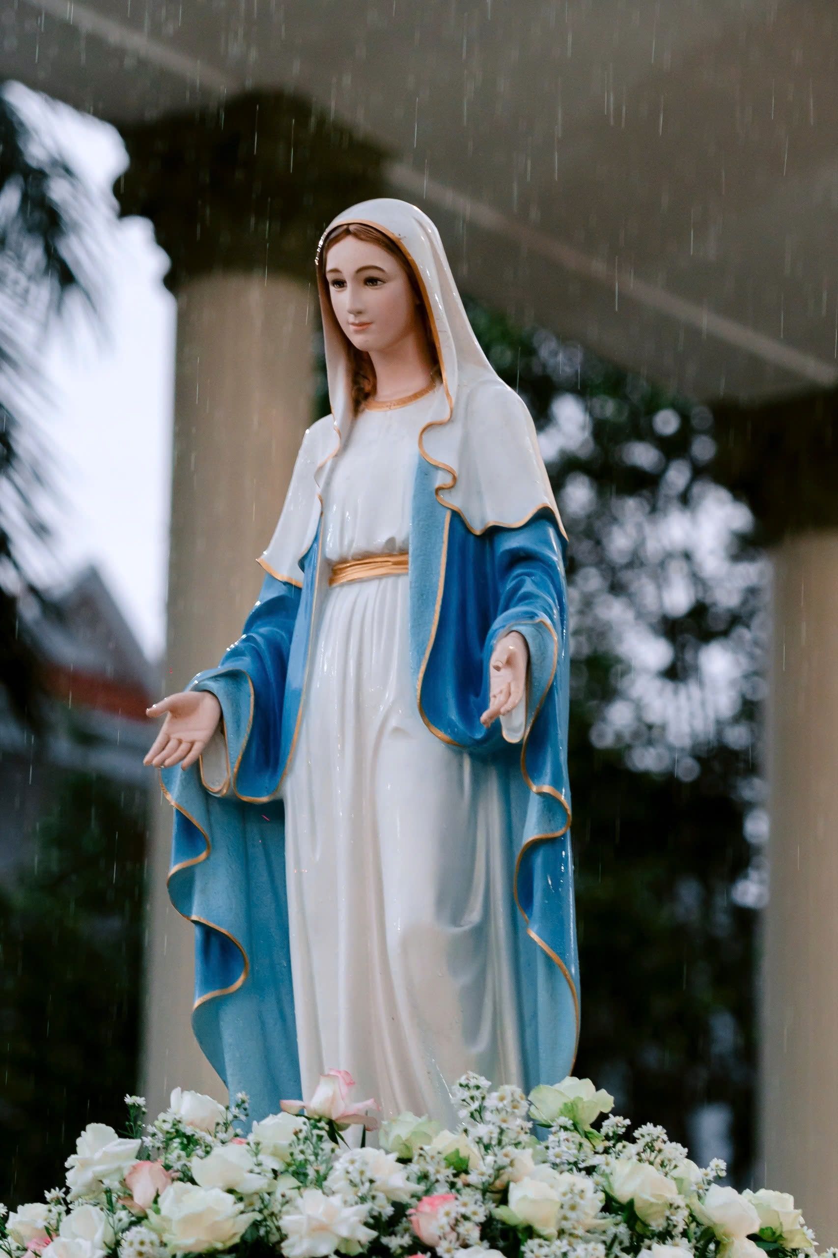 A graceful statue of the Virgin Mary surrounded by flowers in a peaceful outdoor setting.