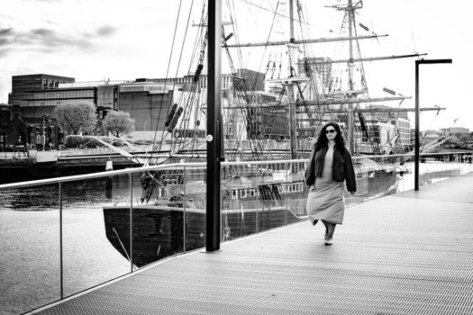Black and white image of a woman walking by tall ships on Dublin's docks, capturing urban maritime life.