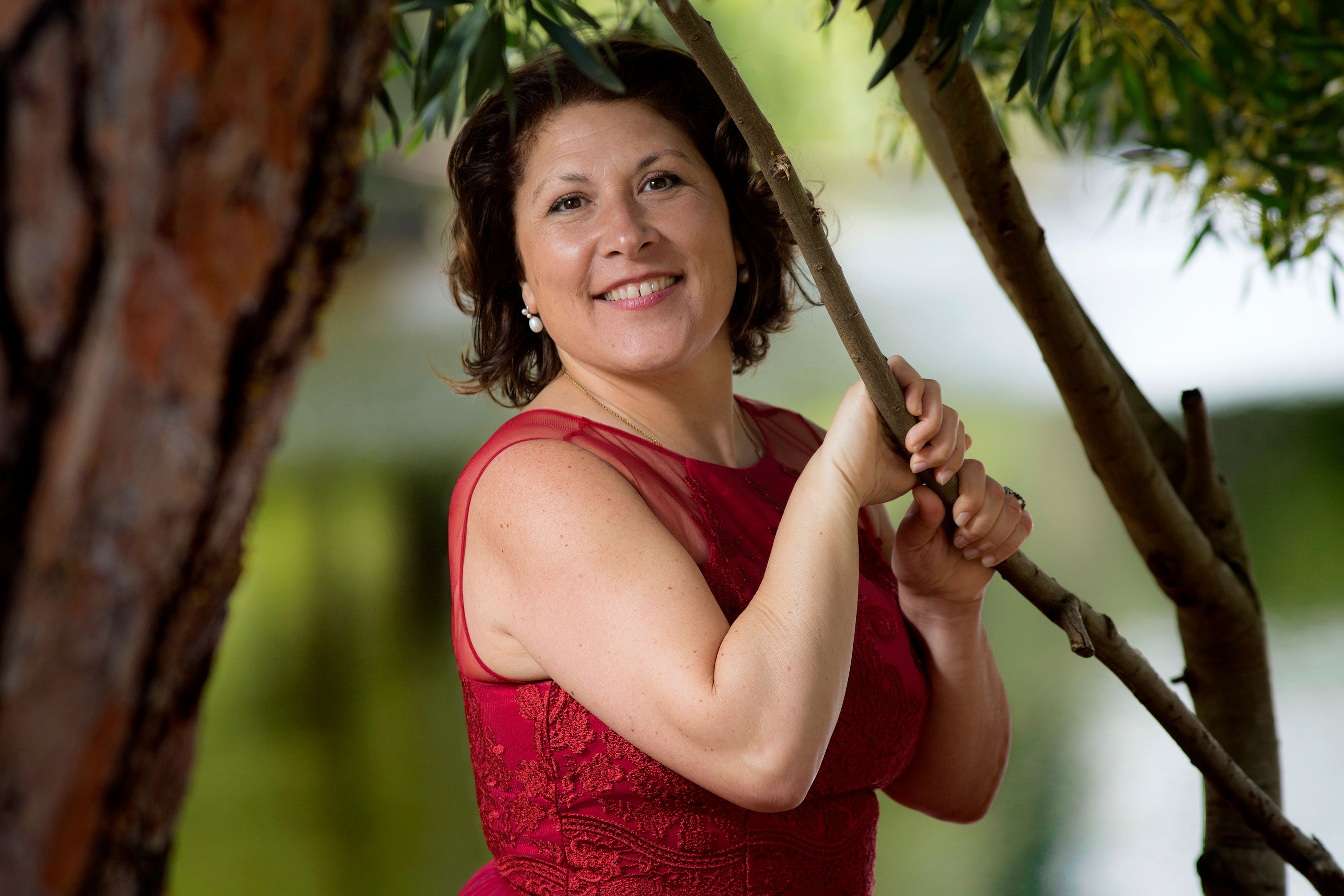 Elegant woman in a red dress smiling and leaning on a tree outdoors.