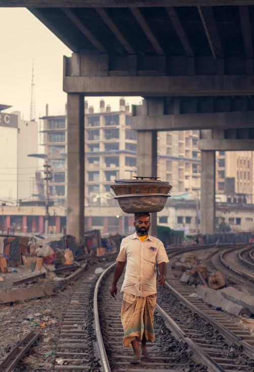 Free Man carrying basket on head, walking along railway tracks under city bridge. Stock Photo