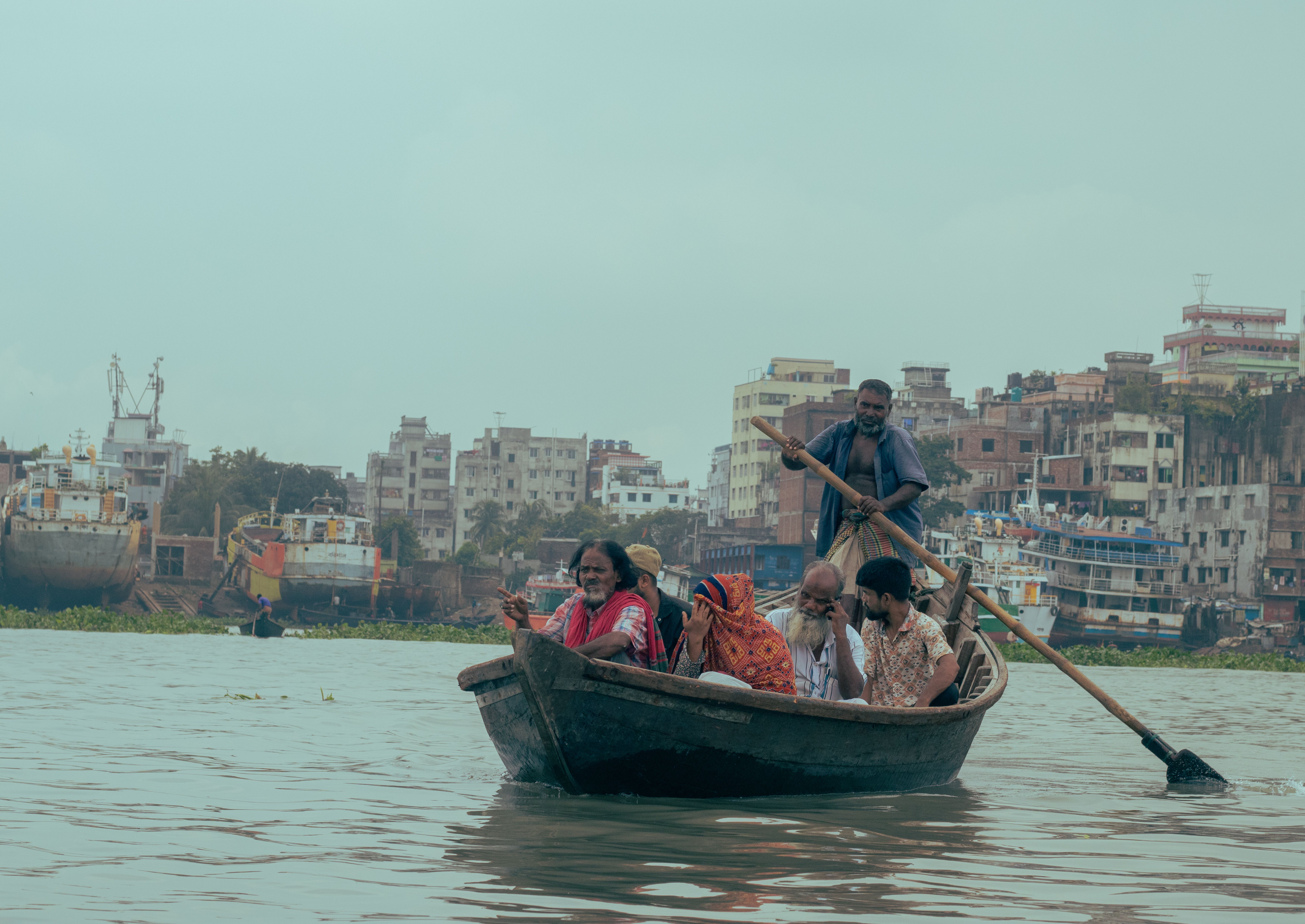 A group of people rowing a boat on a river with a shipyard and urban buildings in the background. — photo by Kaji Rimon / Pexels