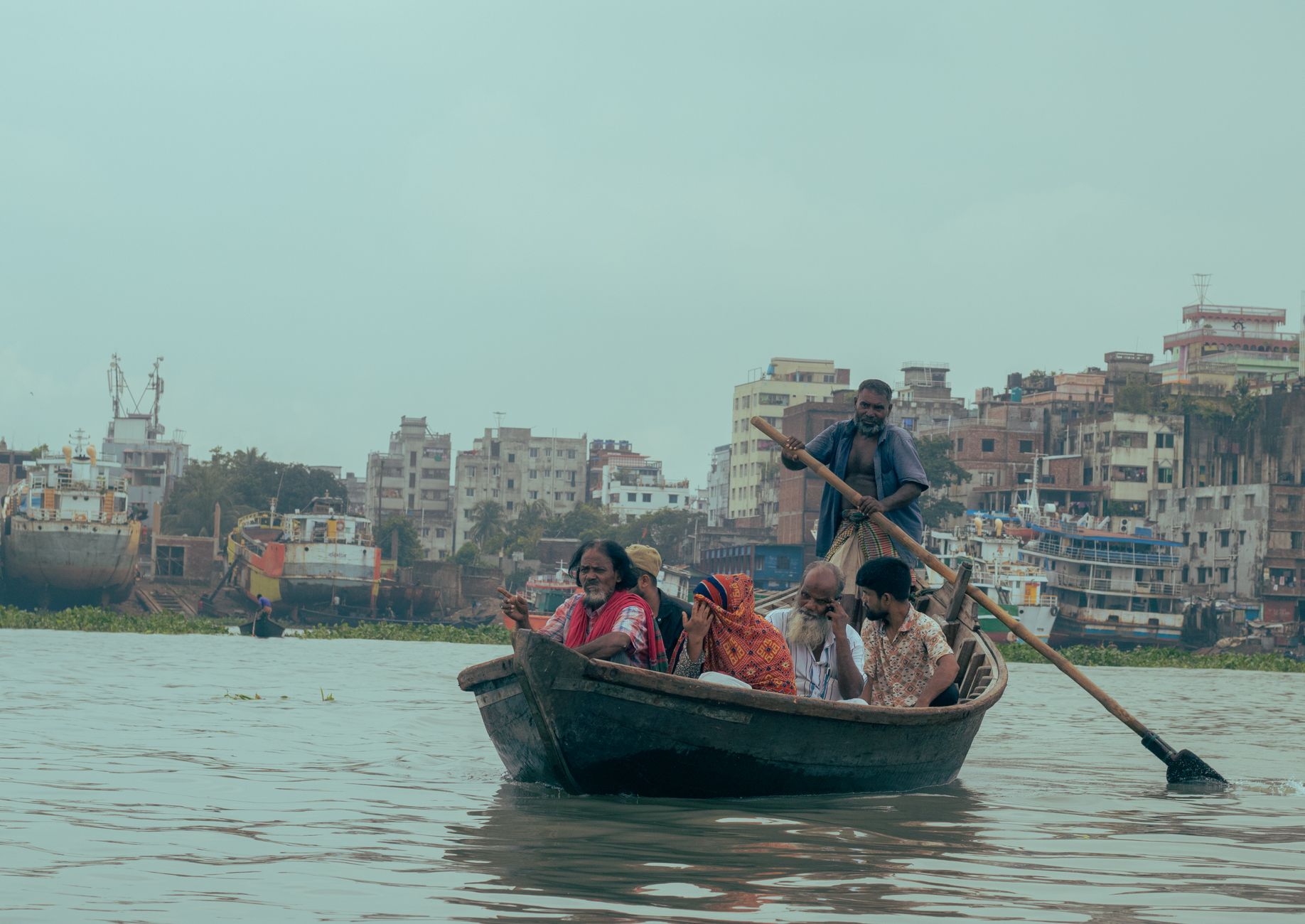 A group of people rowing a boat on a river with a shipyard and urban buildings in the background. โ photo by Kaji Rimon / Pexels