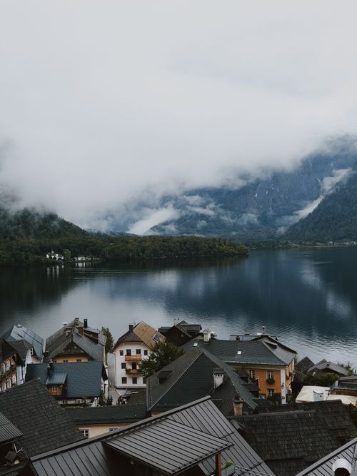 Free Charming lake village nestled in the Alps with misty mountains and serene waters reflecting nature's tranquility. Stock Photo
