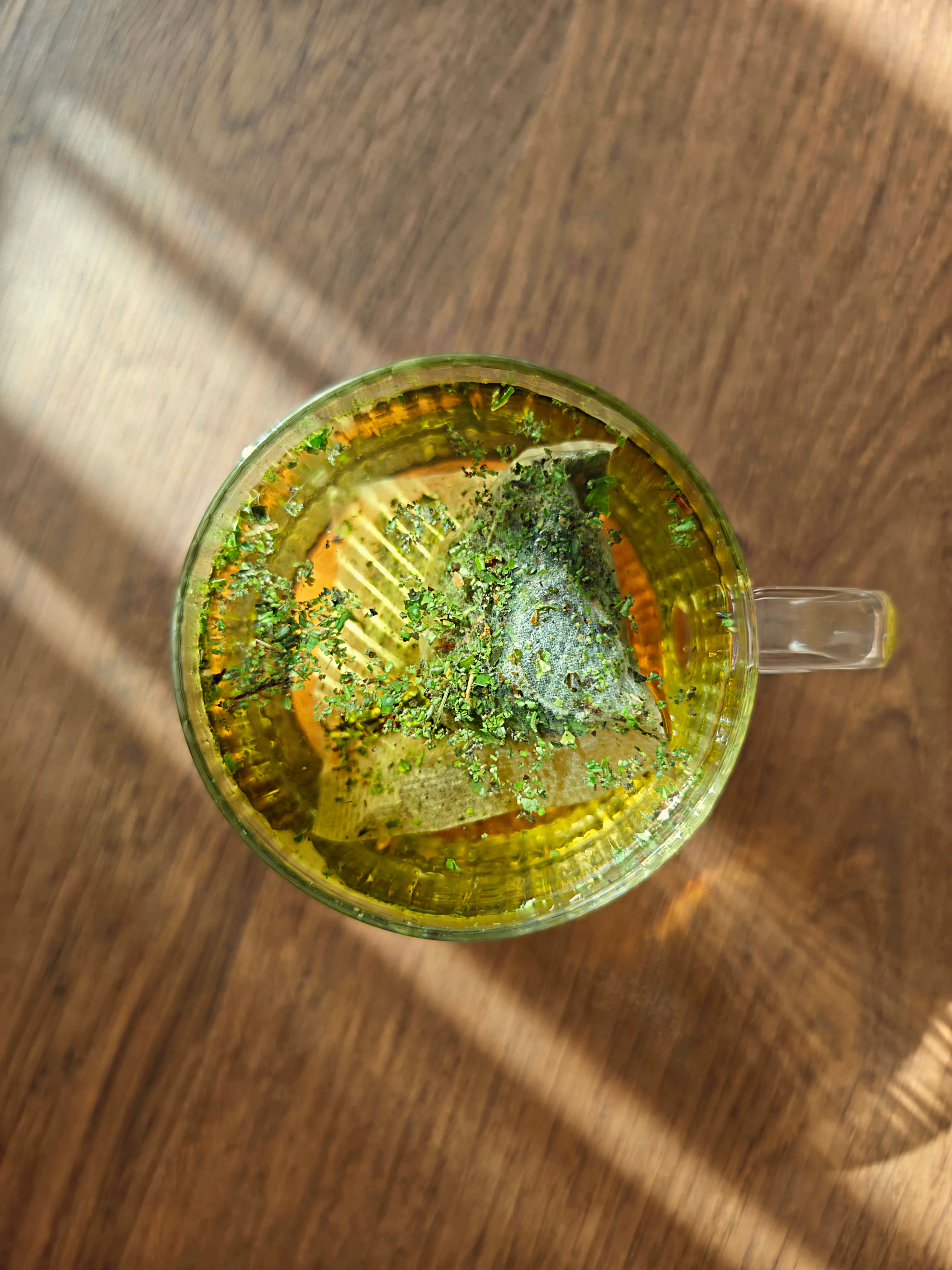 A warm herbal tea with visible herbs in a transparent glass mug on a wooden surface.