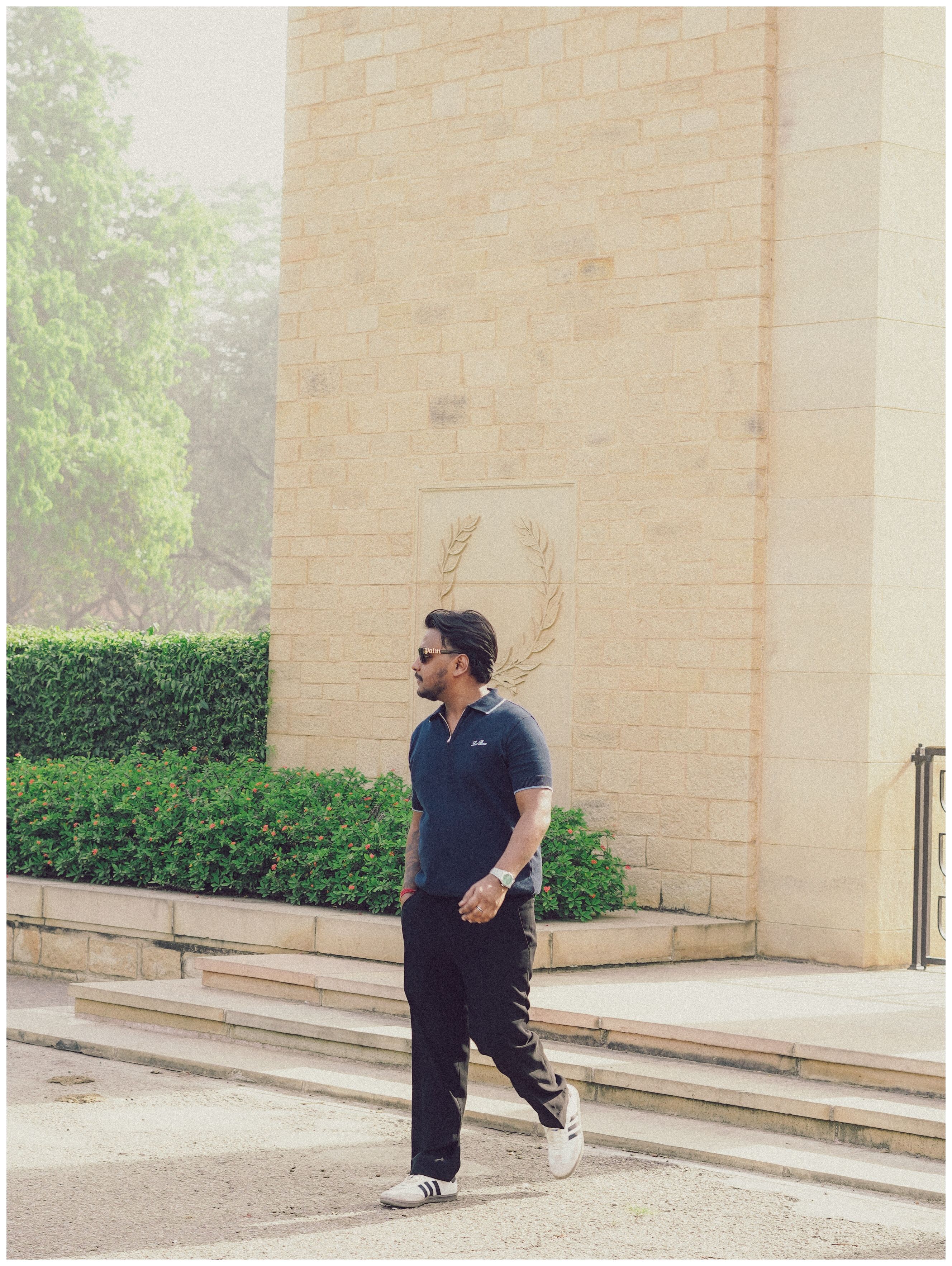 Man walking casually by a stone wall with greenery on a sunny day.
