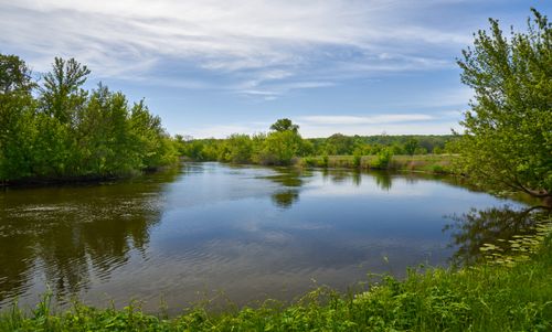 Free A tranquil river scene surrounded by lush greenery and trees reflecting in the water under a bright spring sky. Stock Photo