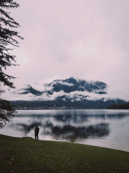 Free A lone figure stands by a foggy mountain lake, reflecting serene wilderness. Stock Photo