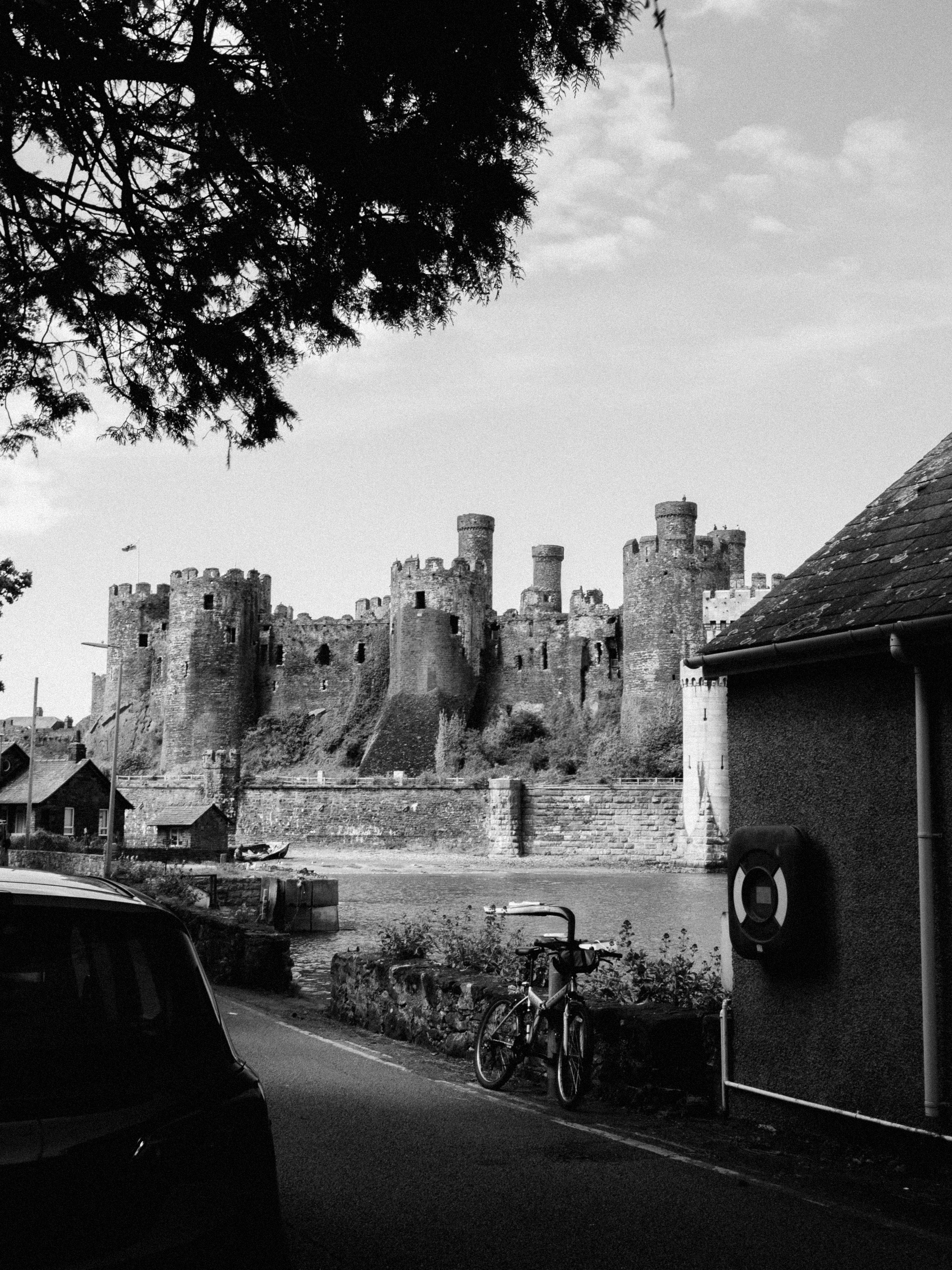 Dramatic black and white photo of the medieval Conwy Castle in Conwy, Wales, with road and bicycle in foreground.