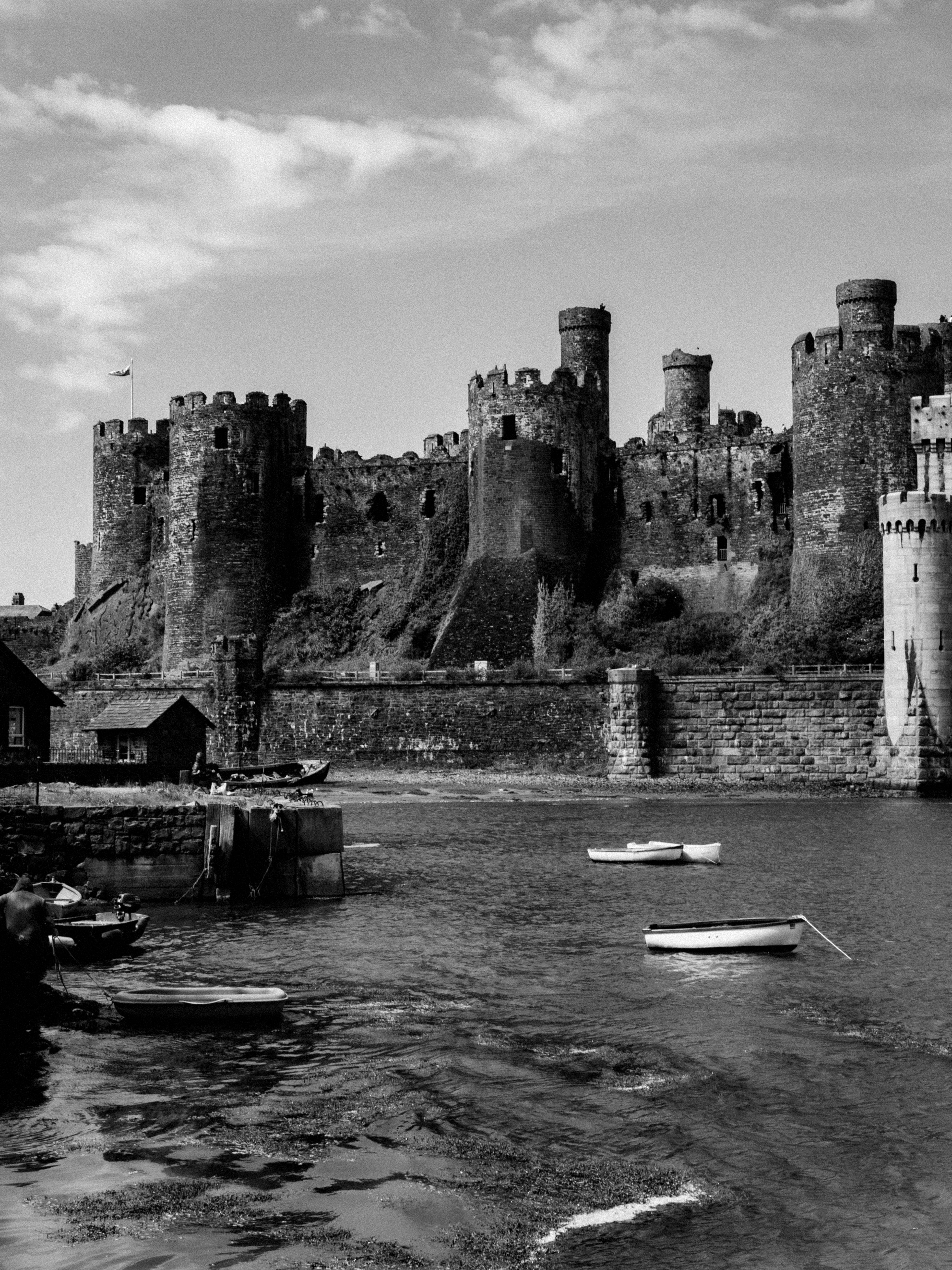 Black and white photograph of Conwy Castle, Wales by the river under a cloudy sky.