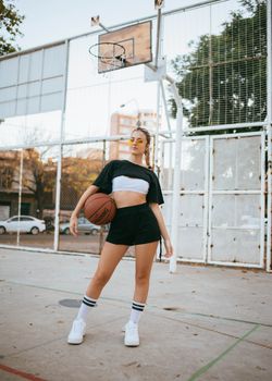 Confident young woman posing with a basketball on an outdoor court, showcasing sporty fashion.