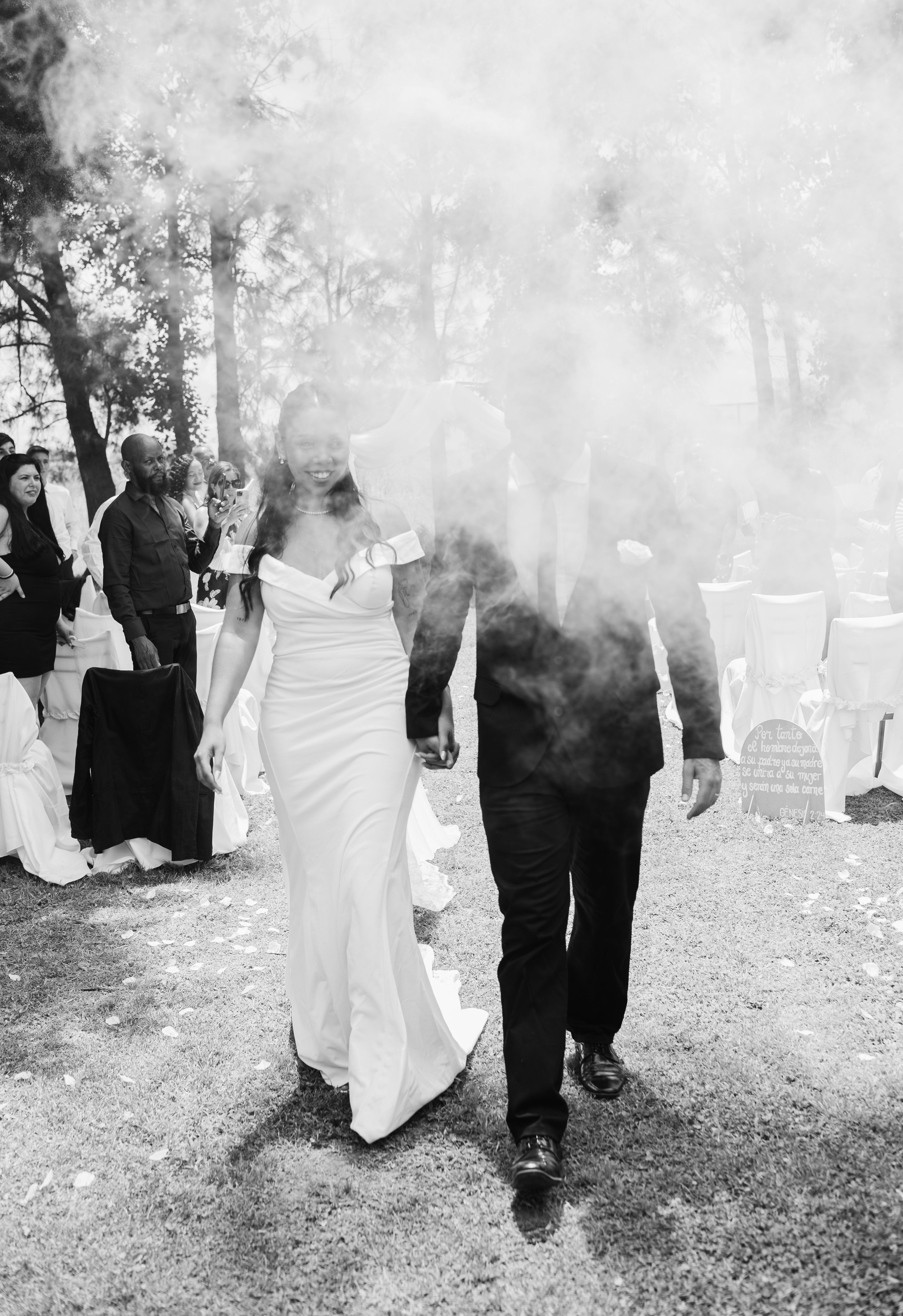 A couple walks through smoke at an outdoor wedding ceremony, captured in black and white.