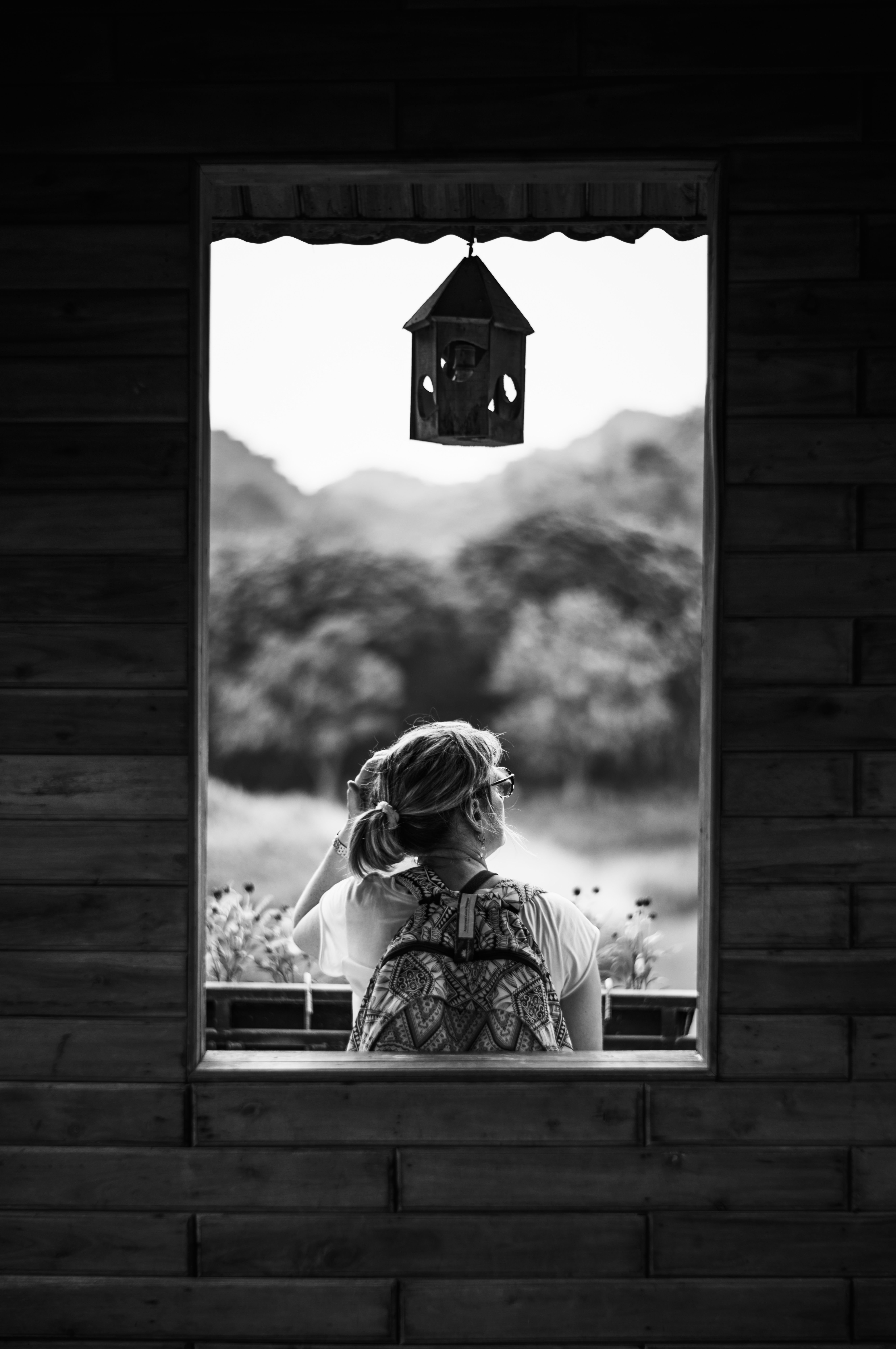 Black and white photo of a woman with backpack gazing through a wooden window in Ninh Bình, Vietnam.