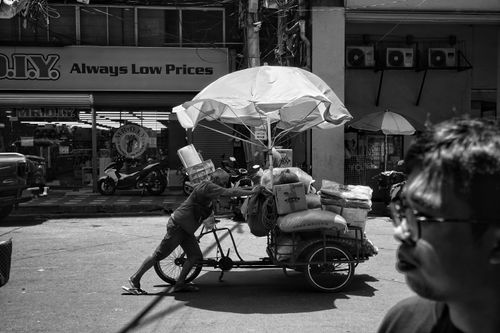 Free Black and white image of a street vendor pushing a bicycle cart with goods in an urban area. Stock Photo