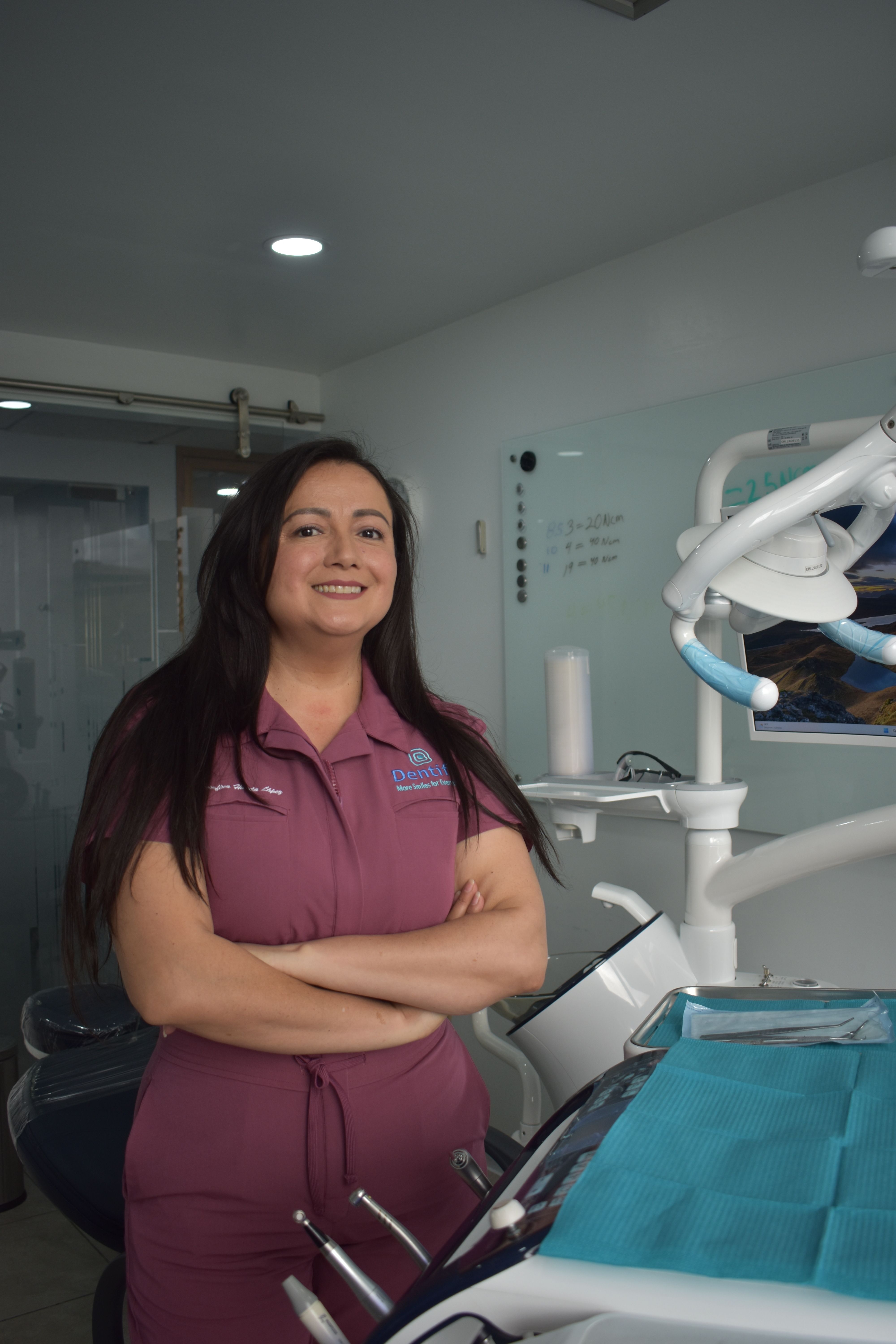 A smiling female dentist stands confidently in a well-equipped dental clinic, ready to treat patients.