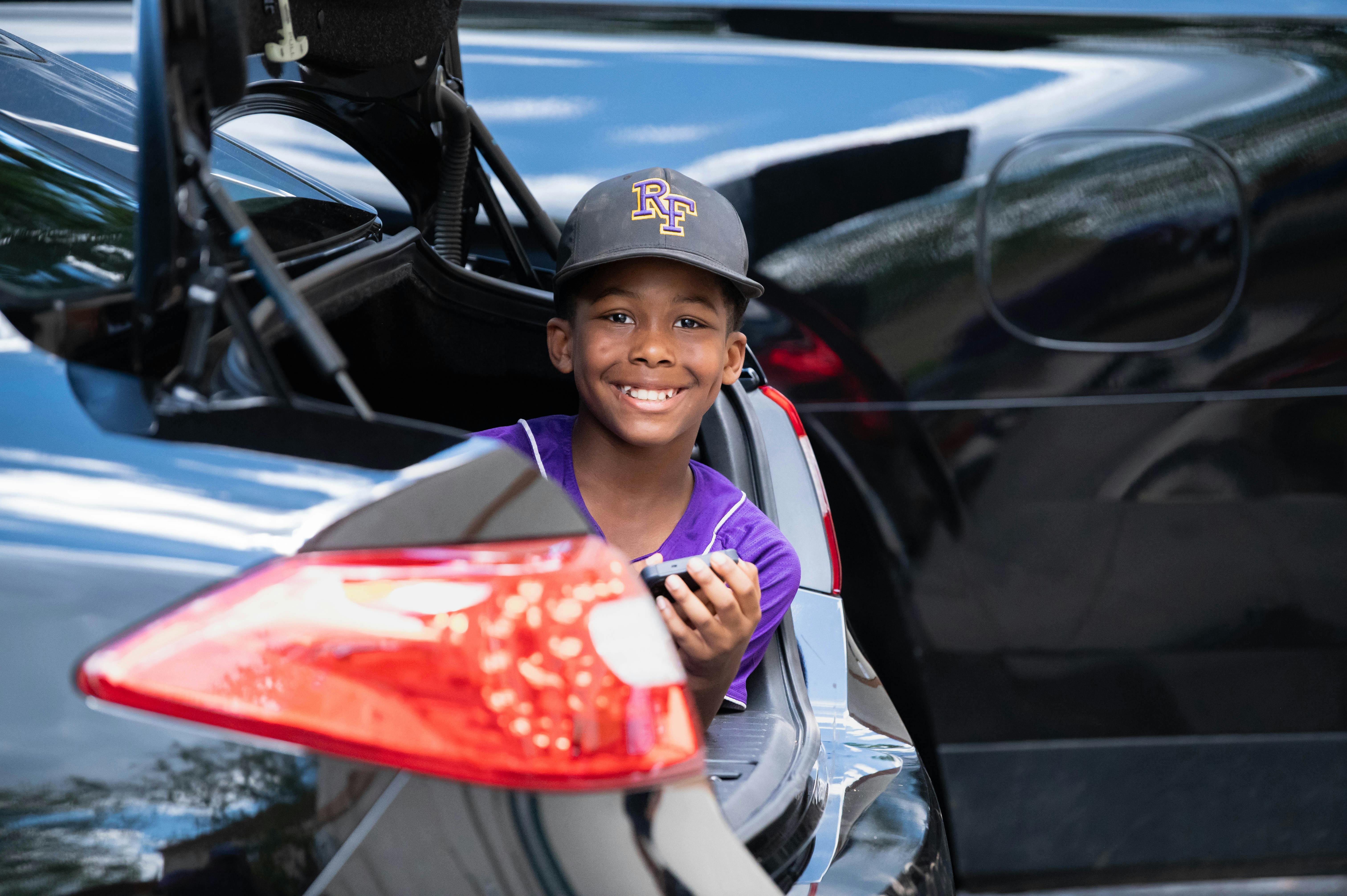 MLB photo featuring Smiling boy inside a car trunk wearing a baseball cap on a bright day, enjoying outdoor playtime.