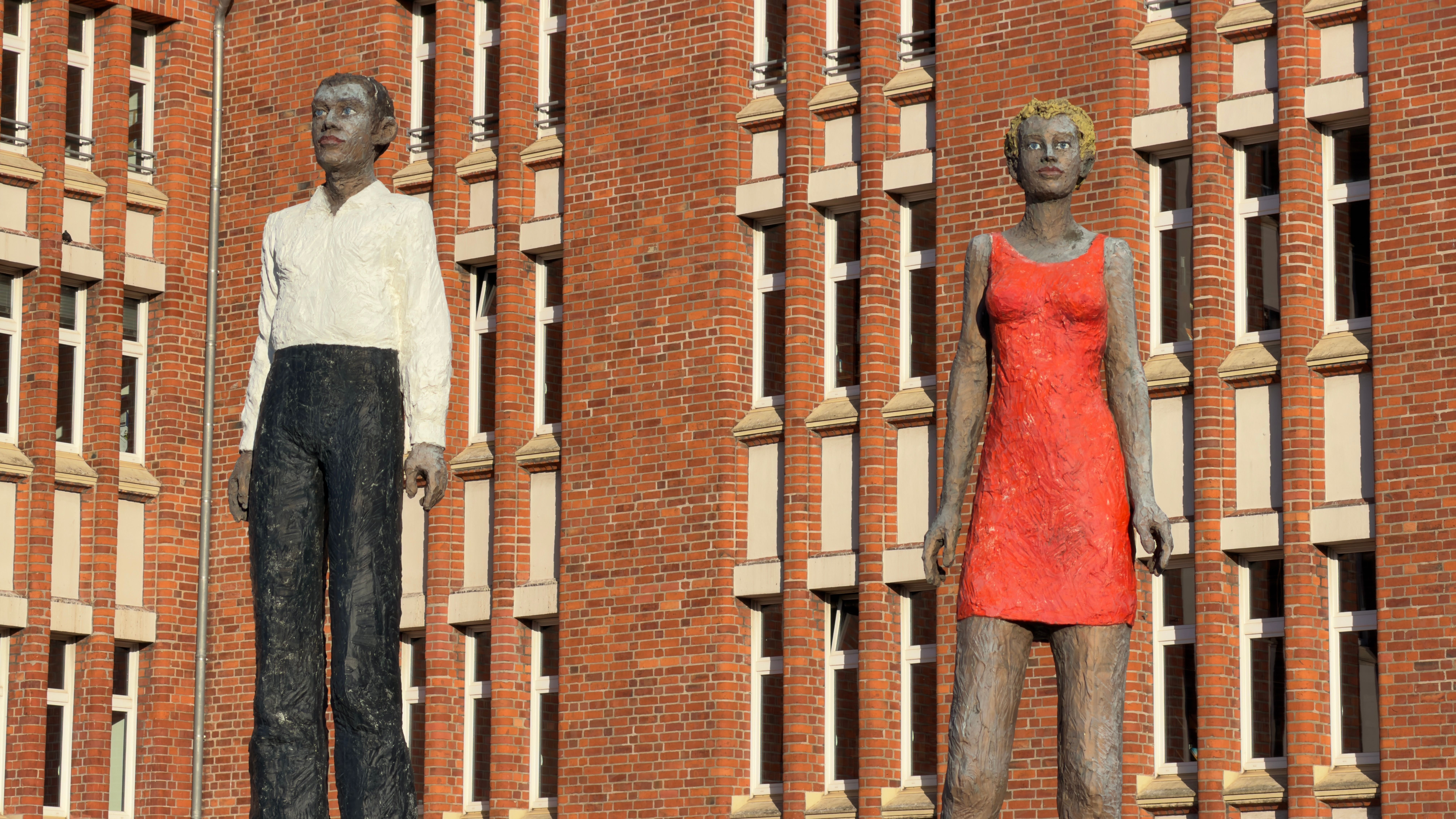 Two tall human sculptures stand prominently against a red brick building facade.