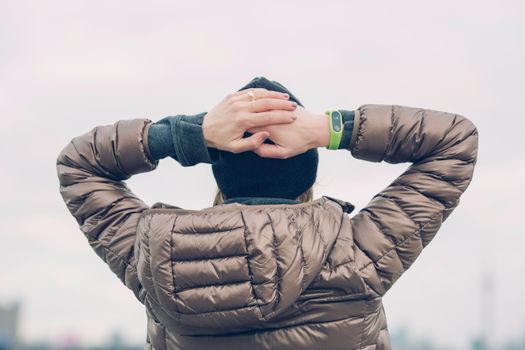 Woman in a winter coat standing outdoors with hands behind head, embracing the cold season.