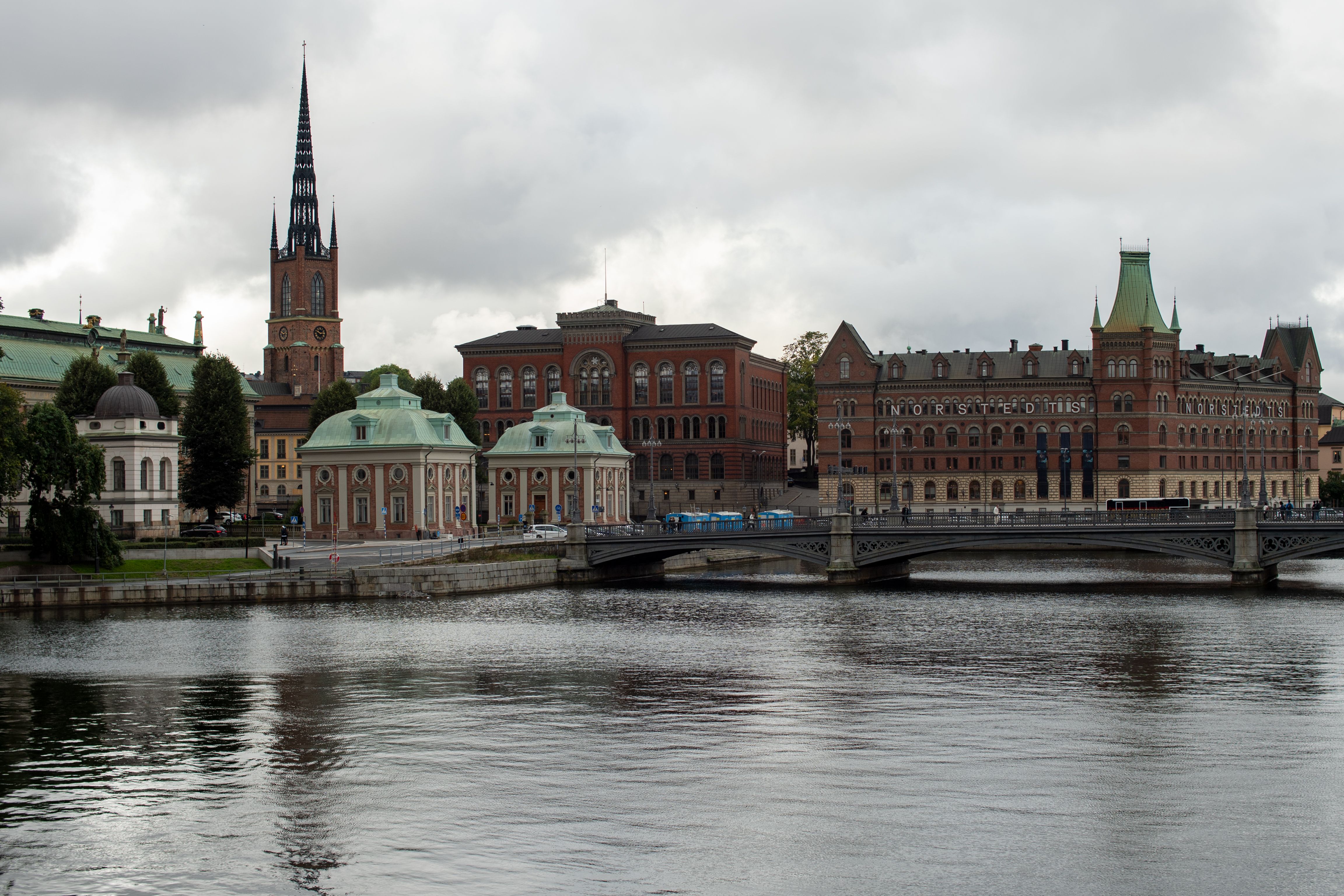 Gratis Utforsk den fredelige utsikten over Stockholms vannkant med den historiske Riddarholmskyrkan og klassisk arkitektur under en stemningsfull himmel. Arkivbilde