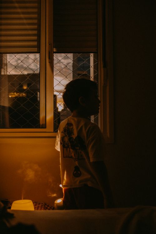 Free A child stands by a window at sunset, cast in warm shadows indoors. Serene and thoughtful moment. Stock Photo
