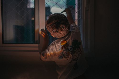 Free A young child gazes out a window in a dimly lit room, wearing a graphic T-shirt. Stock Photo