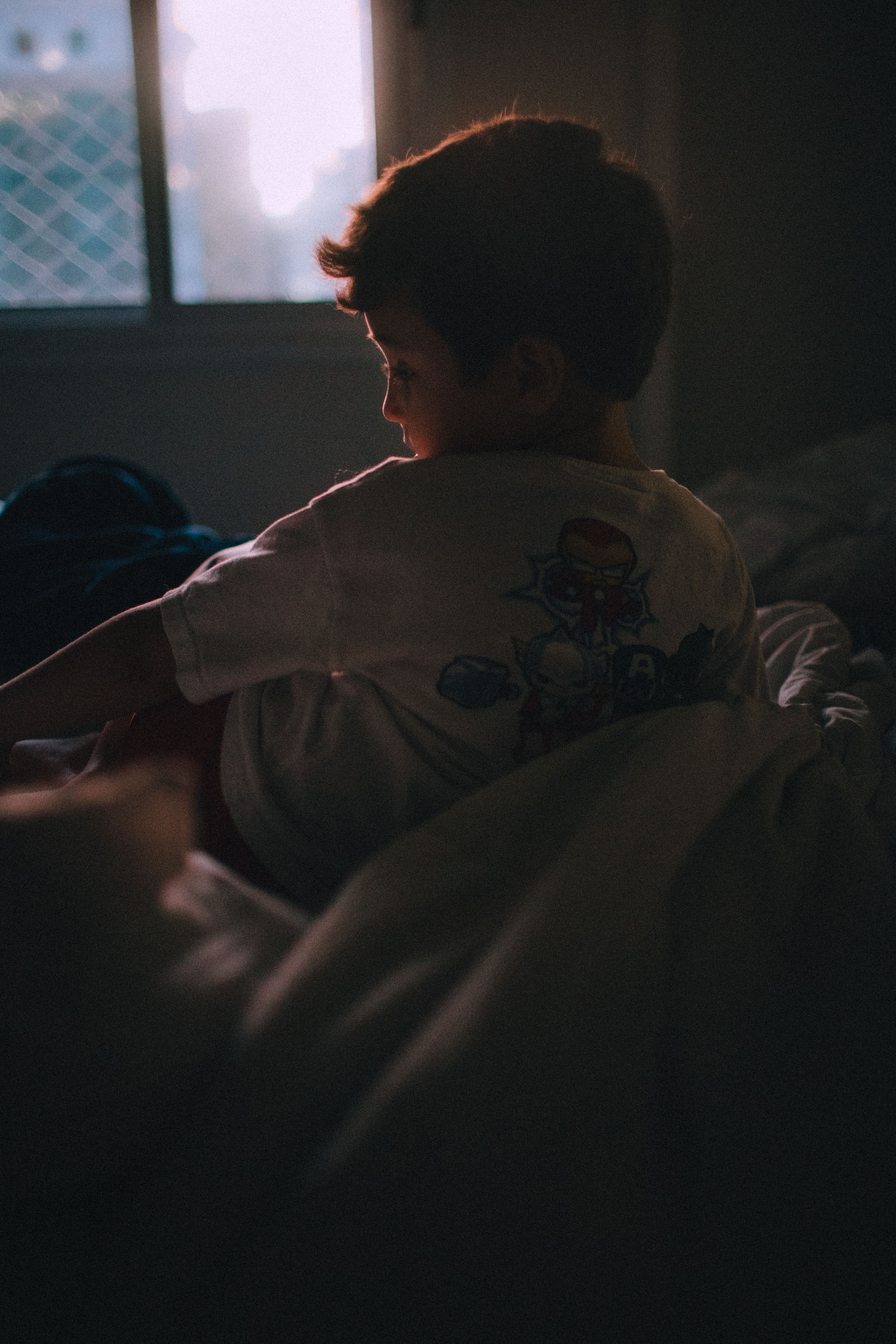 A child gazes thoughtfully at morning light through a window, enveloped in soft shadows.