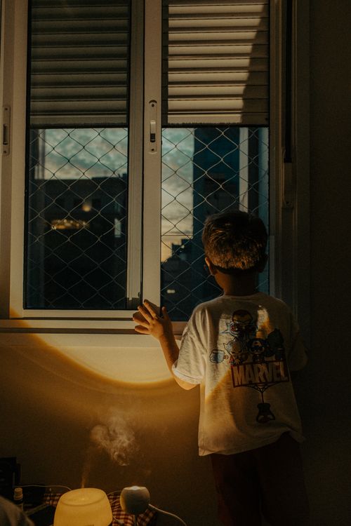 Free A young boy looks out a window at twilight, casting a warm glow indoors. Stock Photo