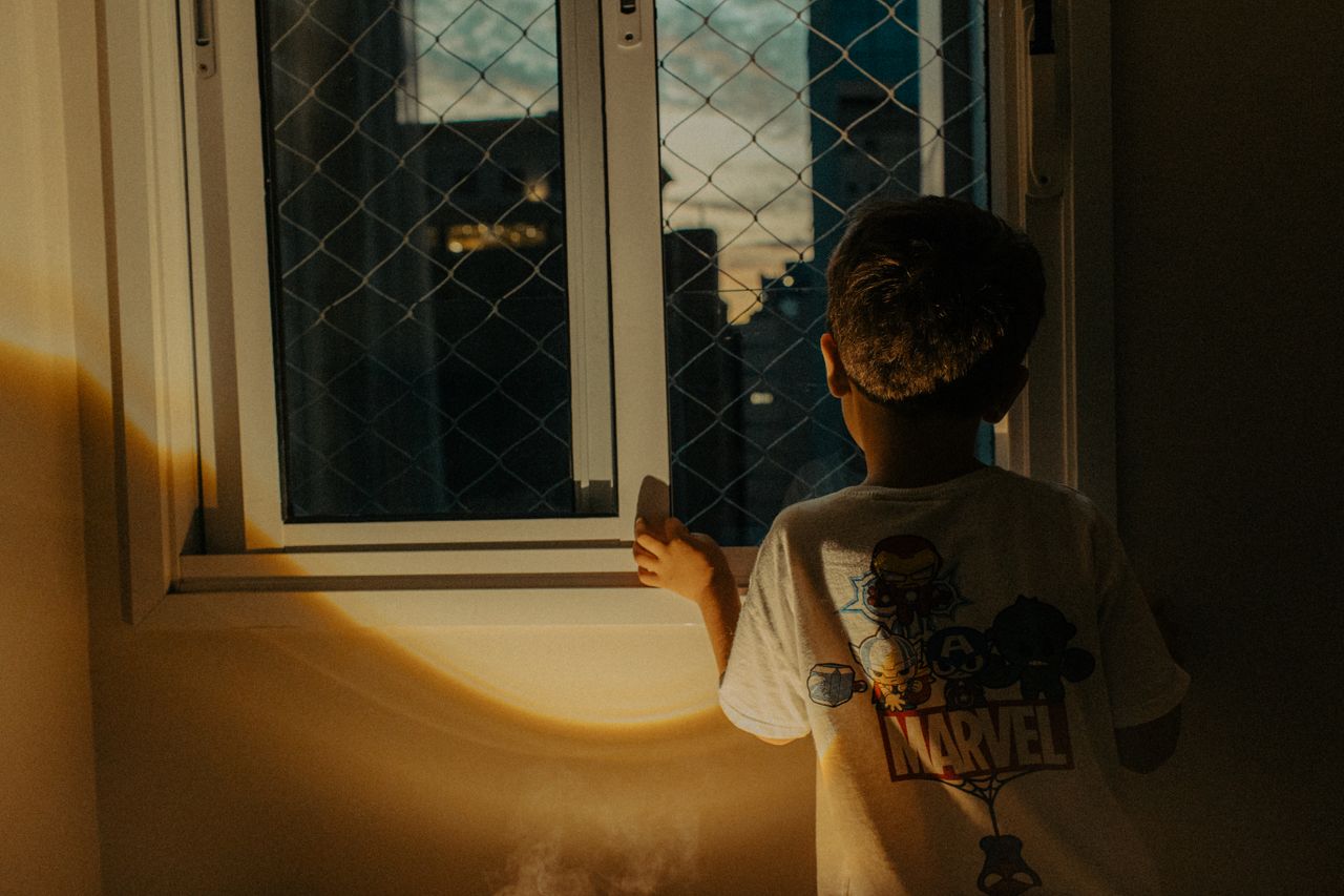 a young boy looks out of a window at sunset wearing a superhero themed