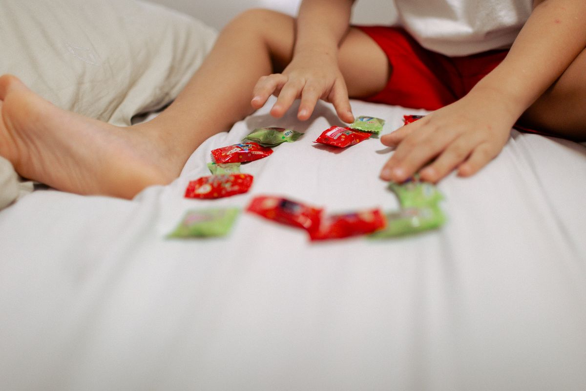 A child arranges colorful candy wrappers on a white bed for a playful indoor scene.