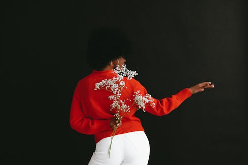 Free Back view of sensual black woman in white denim and white red sweater holding Gypsophila flower behind back posing on black backdrop Stock Photo