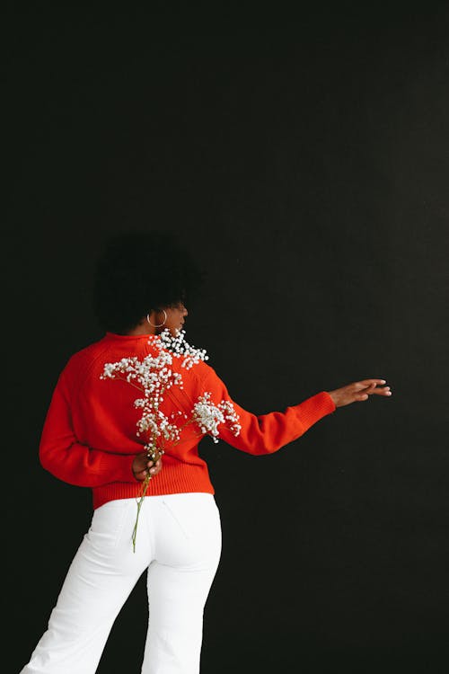 Free Back view of modern African American model wearing vivid red sweater with white bottom holding baby breath flower behind back on black background Stock Photo