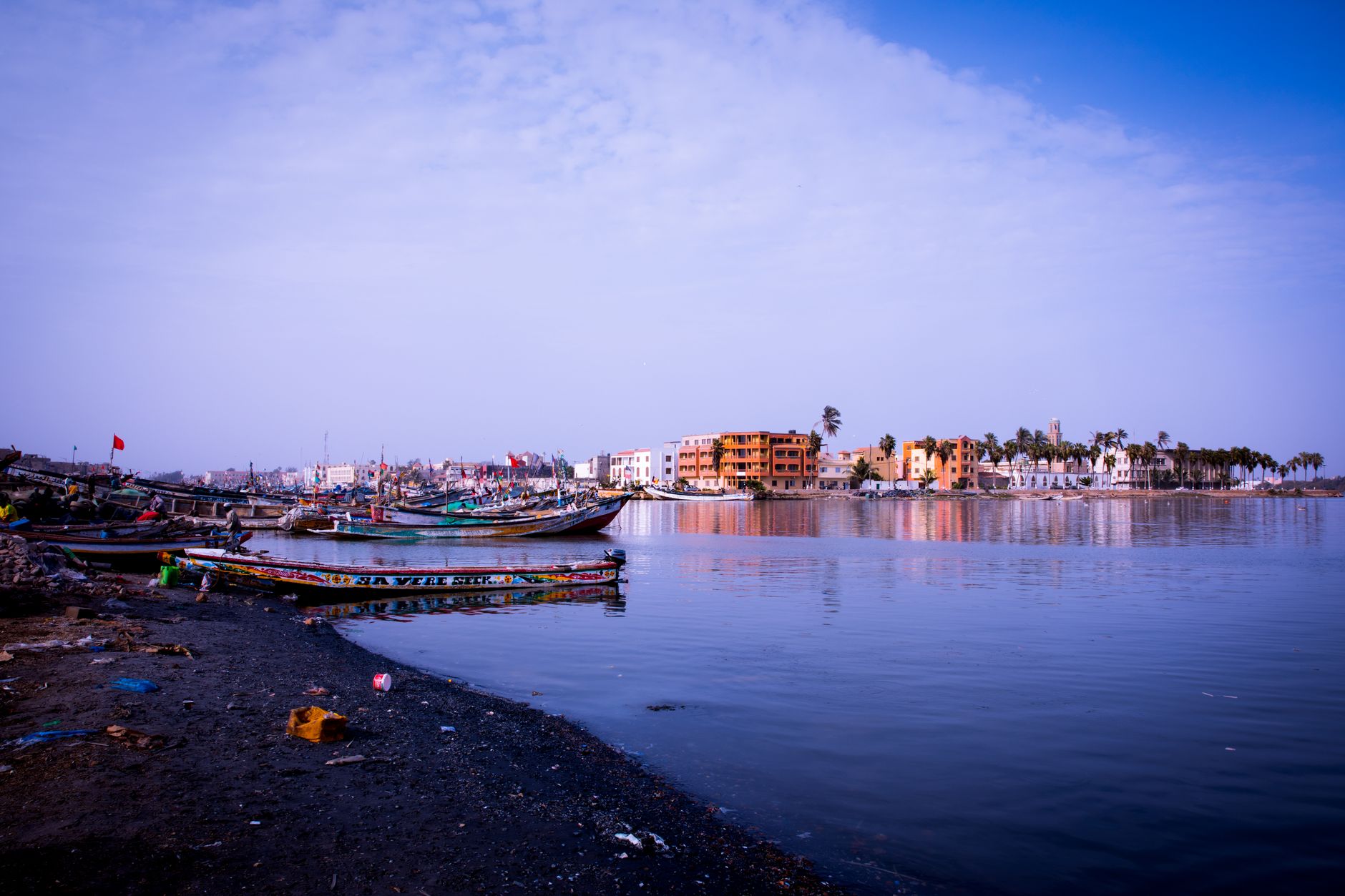 Peaceful coastline scene with moored boats and distant colorful buildings under a clear sky.