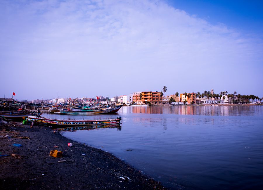 Peaceful coastline scene with moored boats and distant colorful buildings under a clear sky.