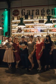 Group of adults in vintage attire posing in a stylish bar with neon lights and retro decor.