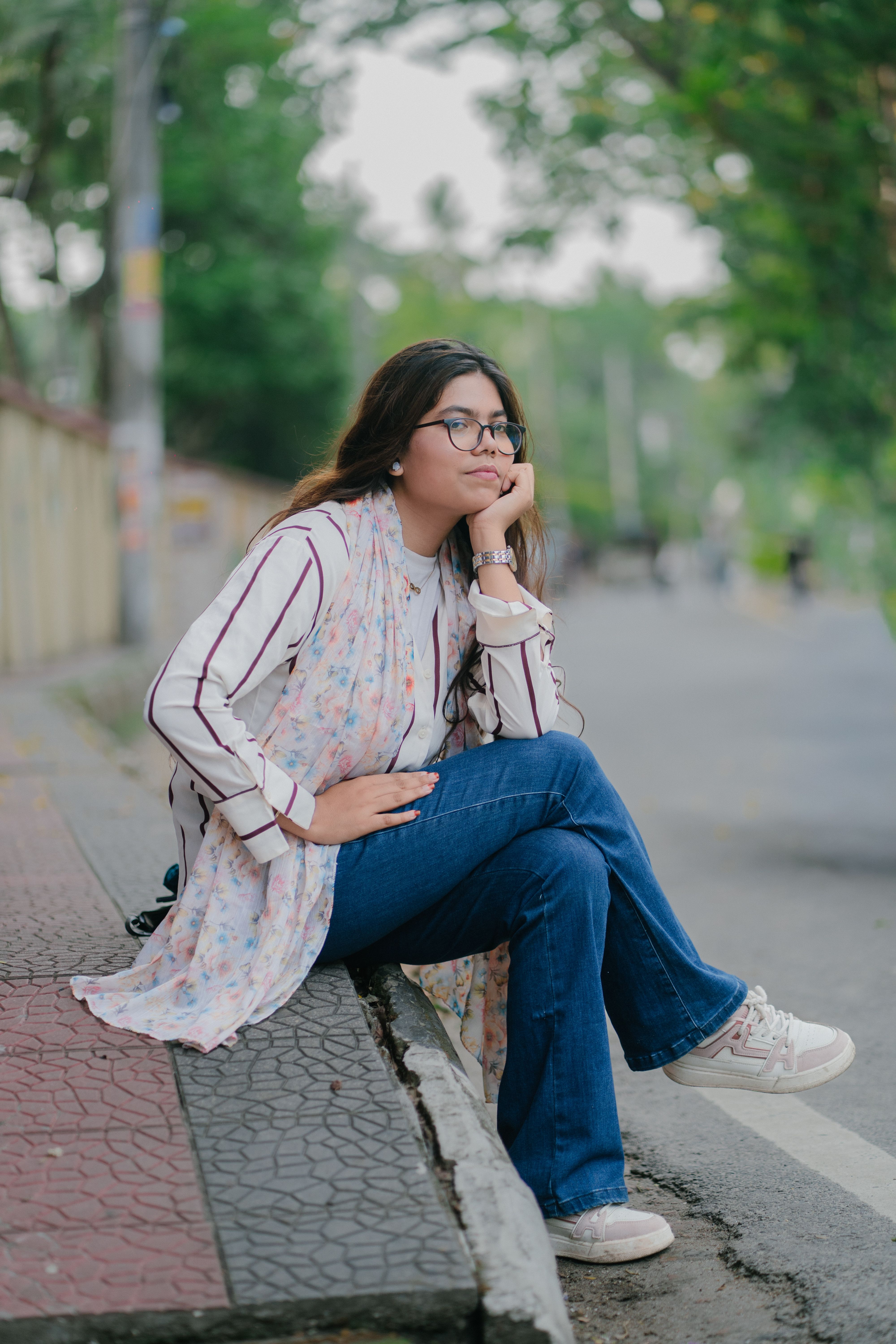 A young woman in casual attire sitting on a sidewalk in a city setting, pondering thoughtfully.
