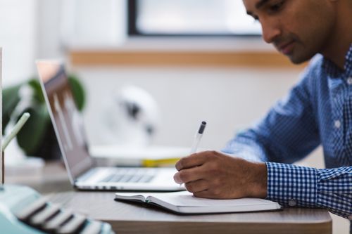Free Man writing in notebook at office desk with laptop, displaying concentration and focus. Stock Photo