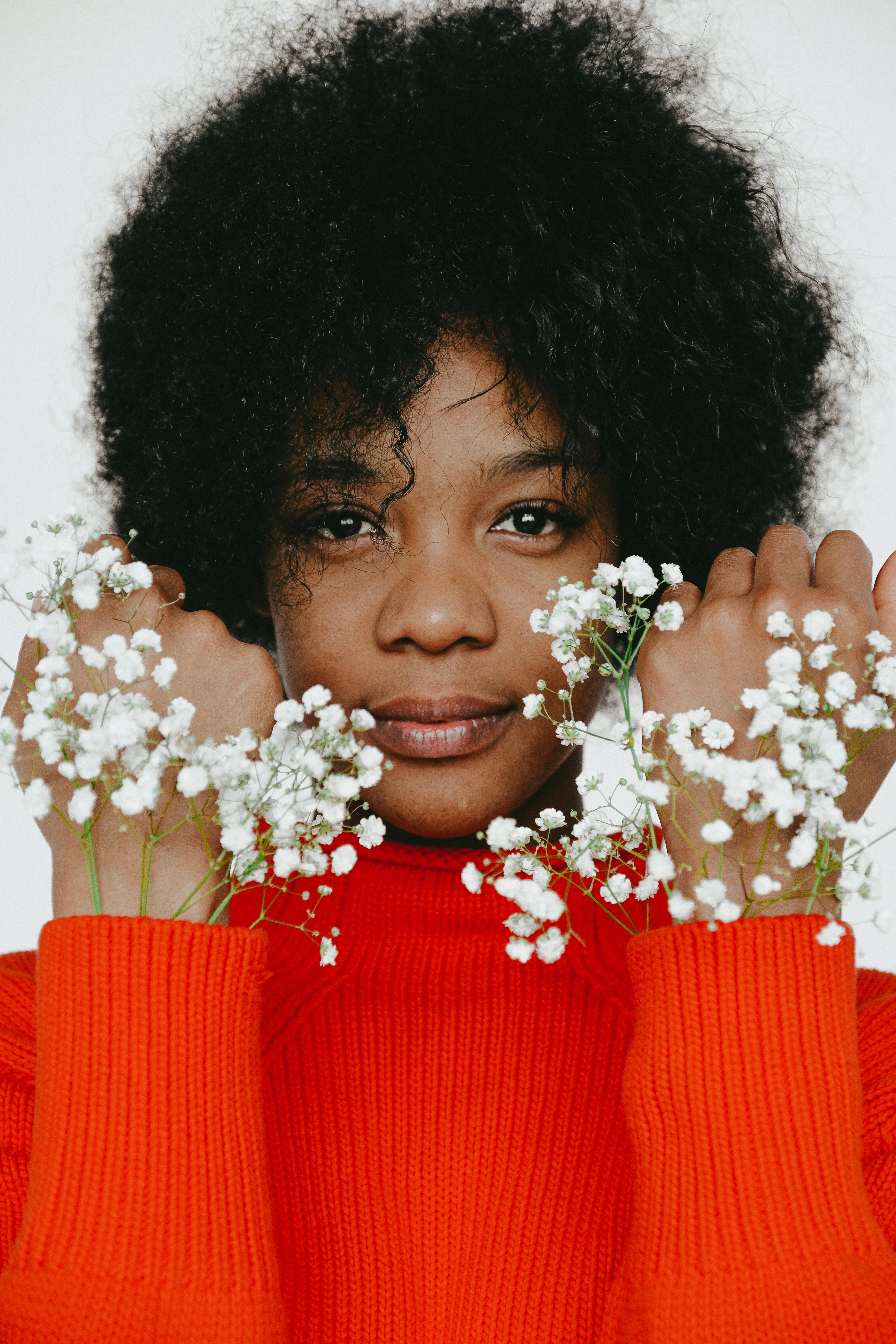 Portrait of a woman with afro hair holding white flowers, wearing a red sweater, against a white background.