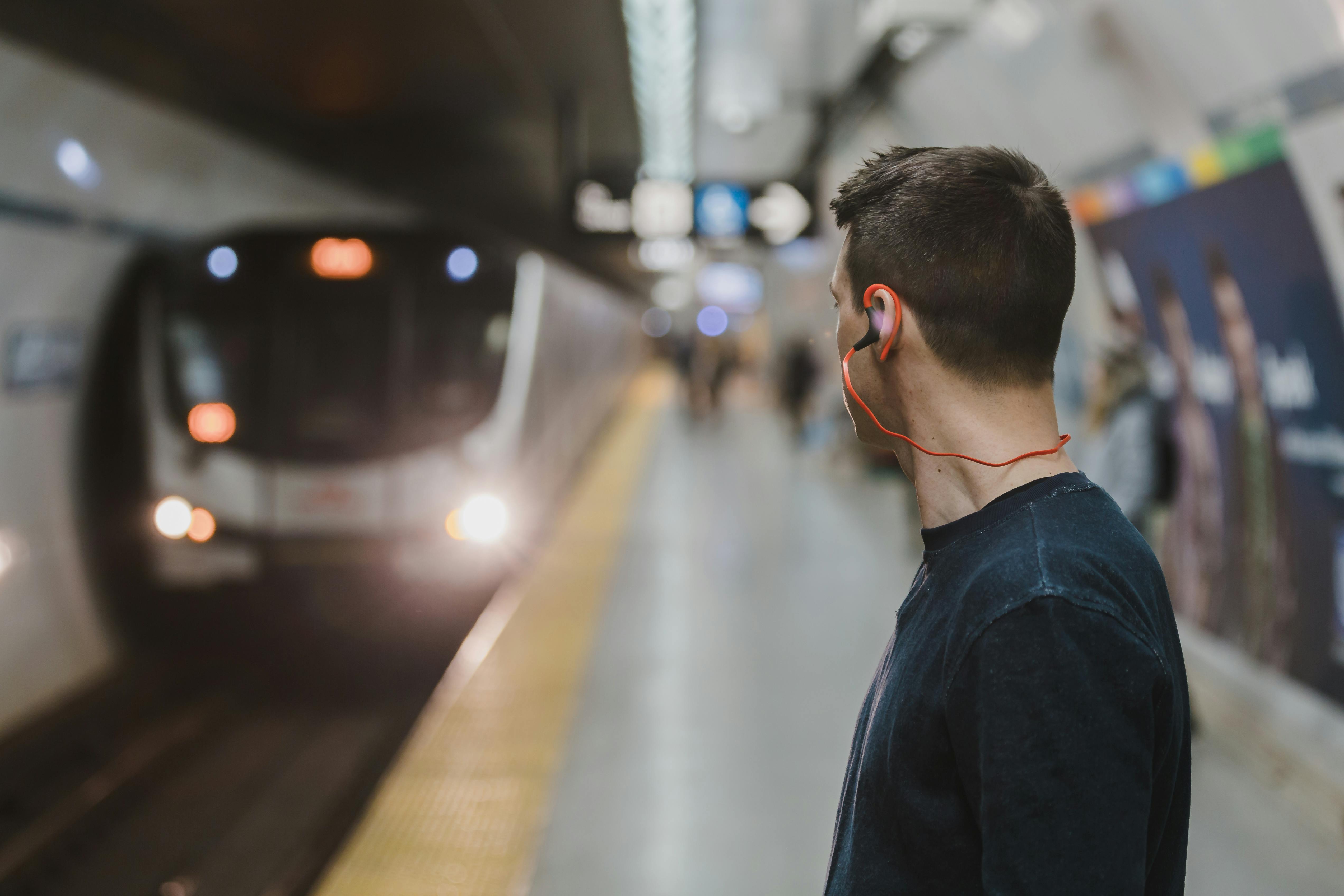 Adult man with earphones waiting on a subway platform, train approaching in urban setting.