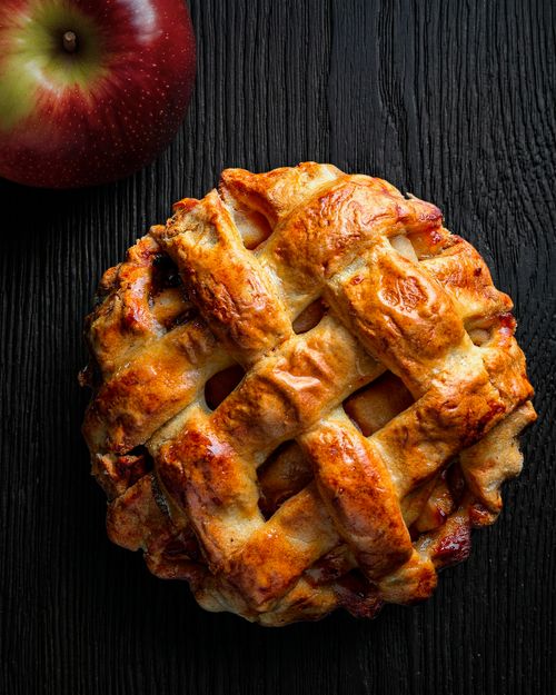 Free A delicious homemade apple pie with a lattice crust on a dark wooden background, accompanied by a red apple. Stock Photo