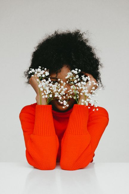 A creative portrait of a woman in a red sweater hiding her face with white flowers against a plain background.