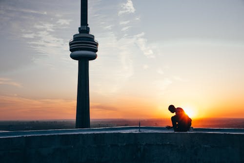 Free Silhouette of a Man Sitting Near Black Tower Near Body of Water during Sunset Stock Photo