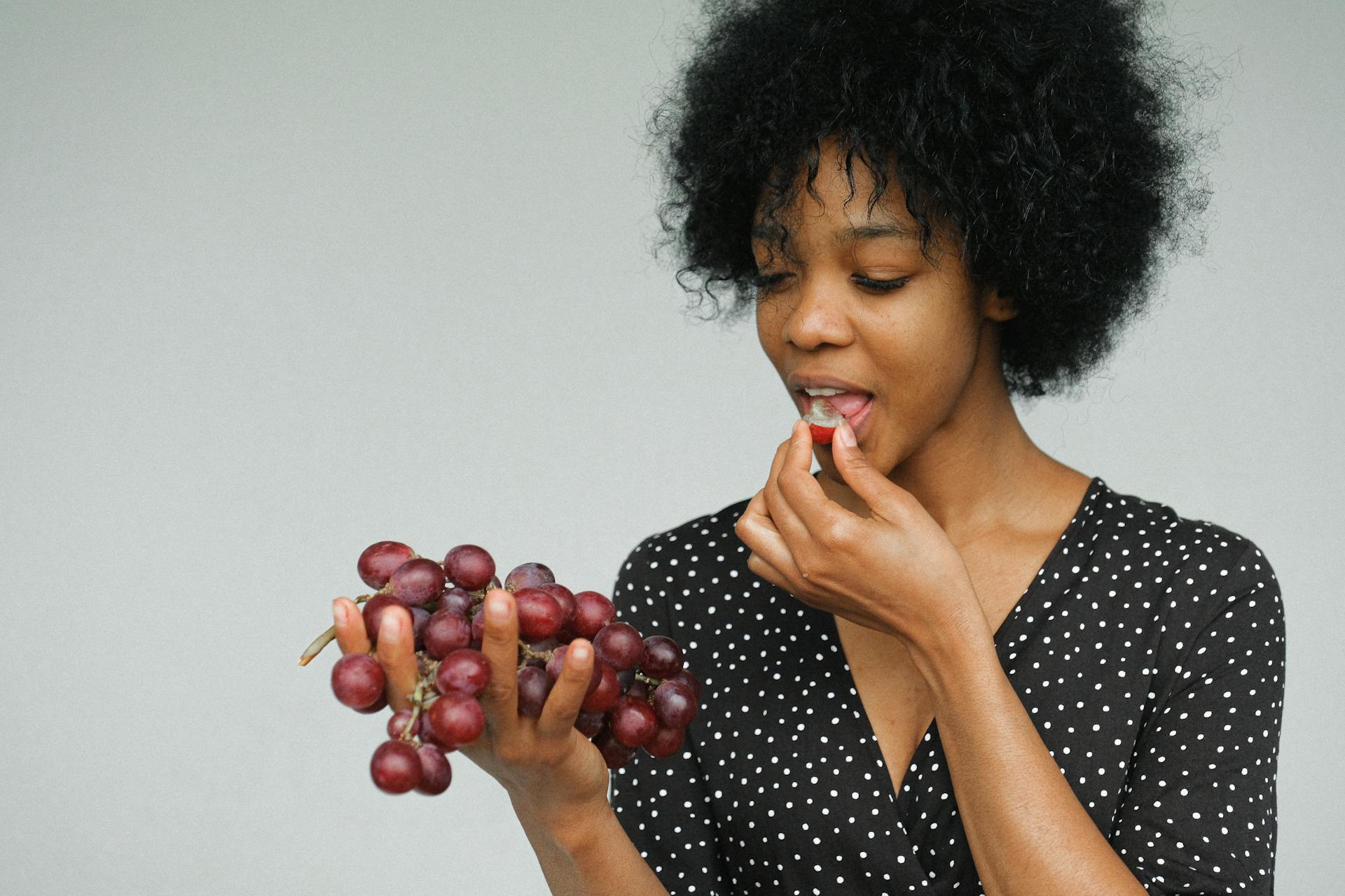 Portrait Photo of Woman in Black and White Polka Dot Dress Eating Grapes While Standing In Front of Gray Background