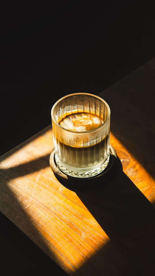 Free A glass of coffee with cream sits on a wooden table, bathed in warm sunlight and surrounded by dark shadows. Stock Photo
