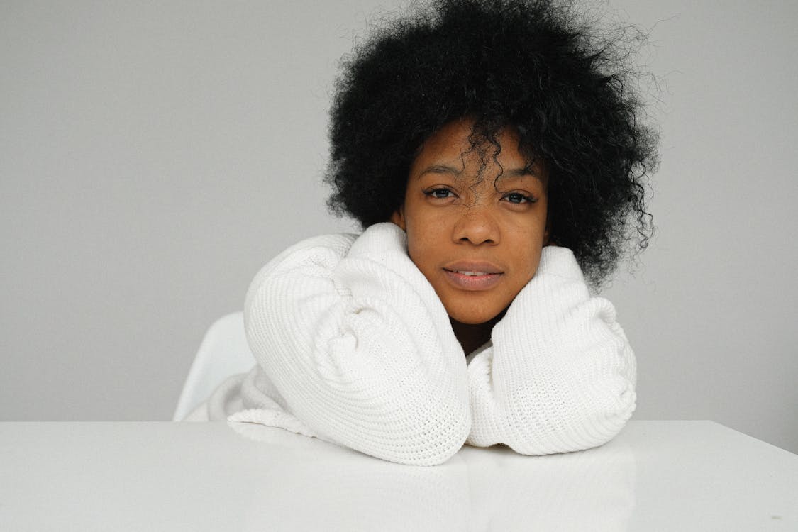Free Charming African American female in white sweater sitting at table and cuddling while looking  at camera on on gray background Stock Photo